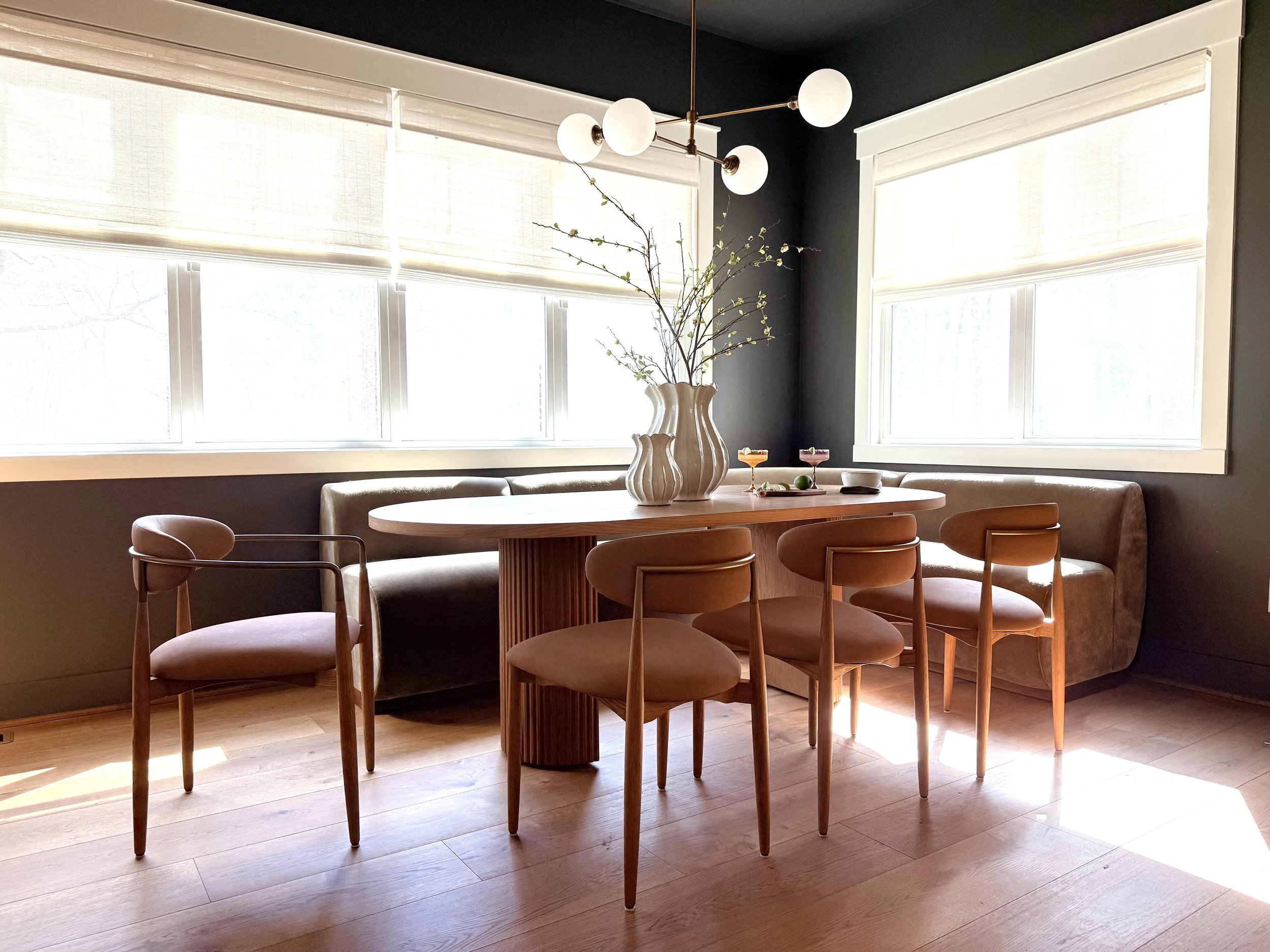 A modern organic dining nook with an oval white oak fluted table and olive-green velvet banquette. Features four mid-century modern chairs with leather seats, a brass globe light fixture, large bright windows, and dark taupe gray contrast walls.