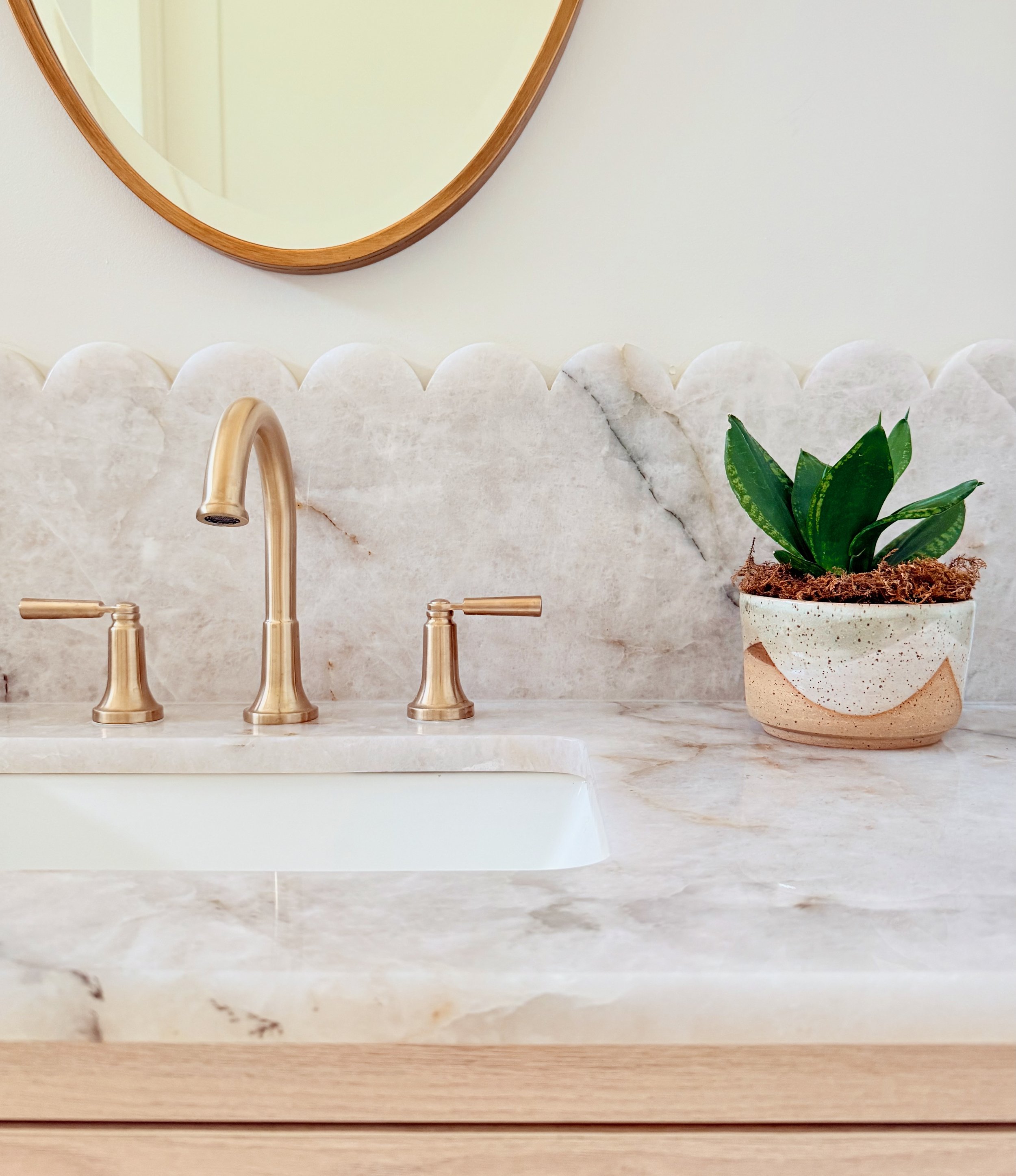 Bathroom sink with a gold faucet and handles, a potted green plant, and a round mirror above