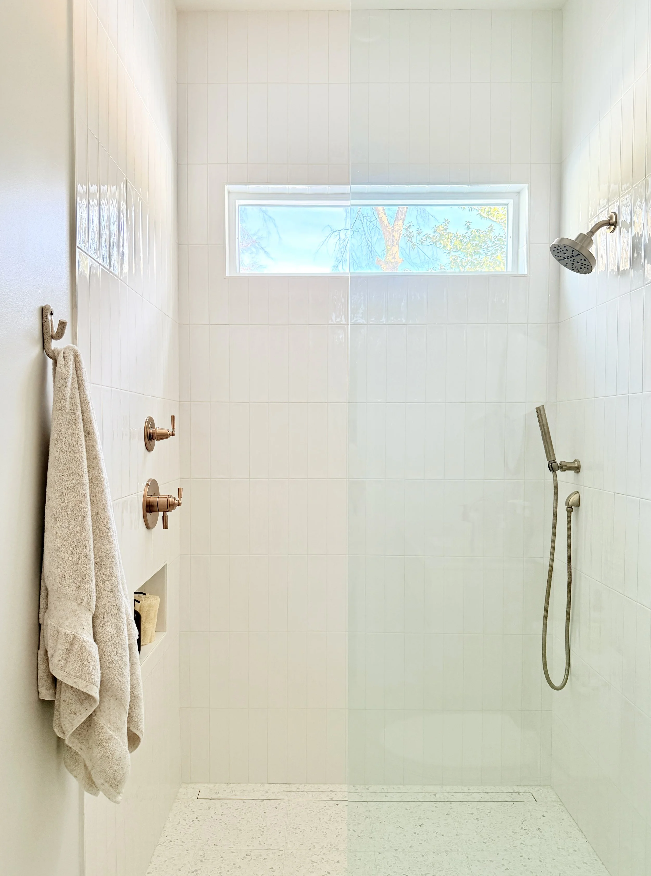 Modern, minimalist shower with white tiled walls, small rectangular window, and showerhead on the right wall. A towel hangs on a hook on the left wall.