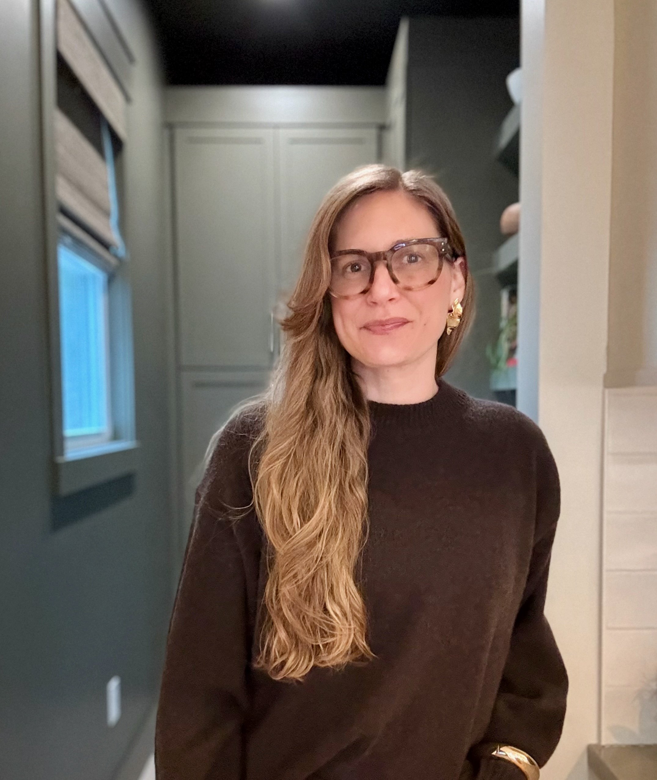 Principal portrait featuring warm, layered style. Wearing tortoise shell glasses and a dark brown sweater, she stands in front of an organic modern pantry with deep green cabinetry.