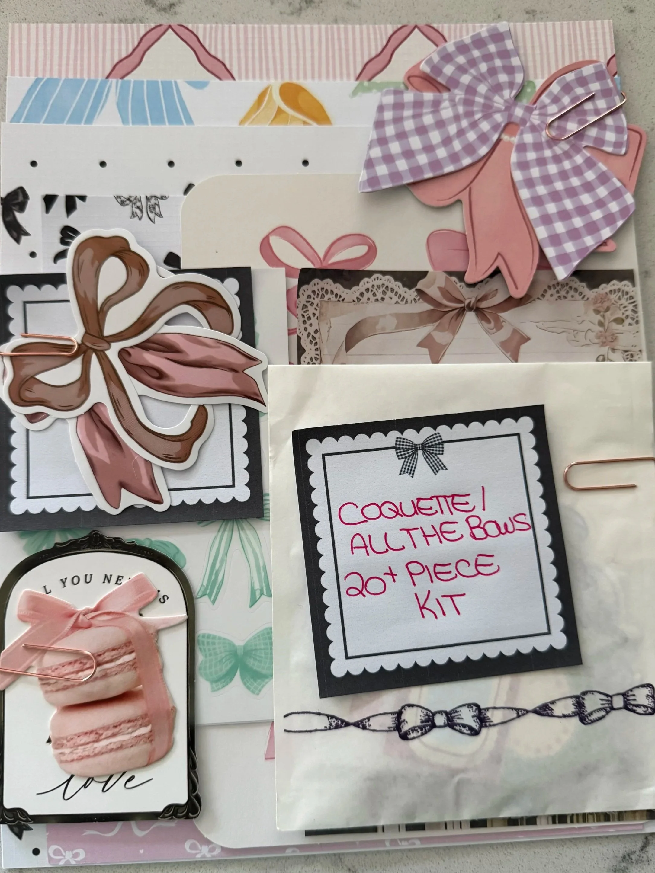 Showcasing a curated journal kit with decorative bow ephemera, patterned papers, paper clips, and a note labeled “Coquette All the Bows 20+ Piece Kit.”