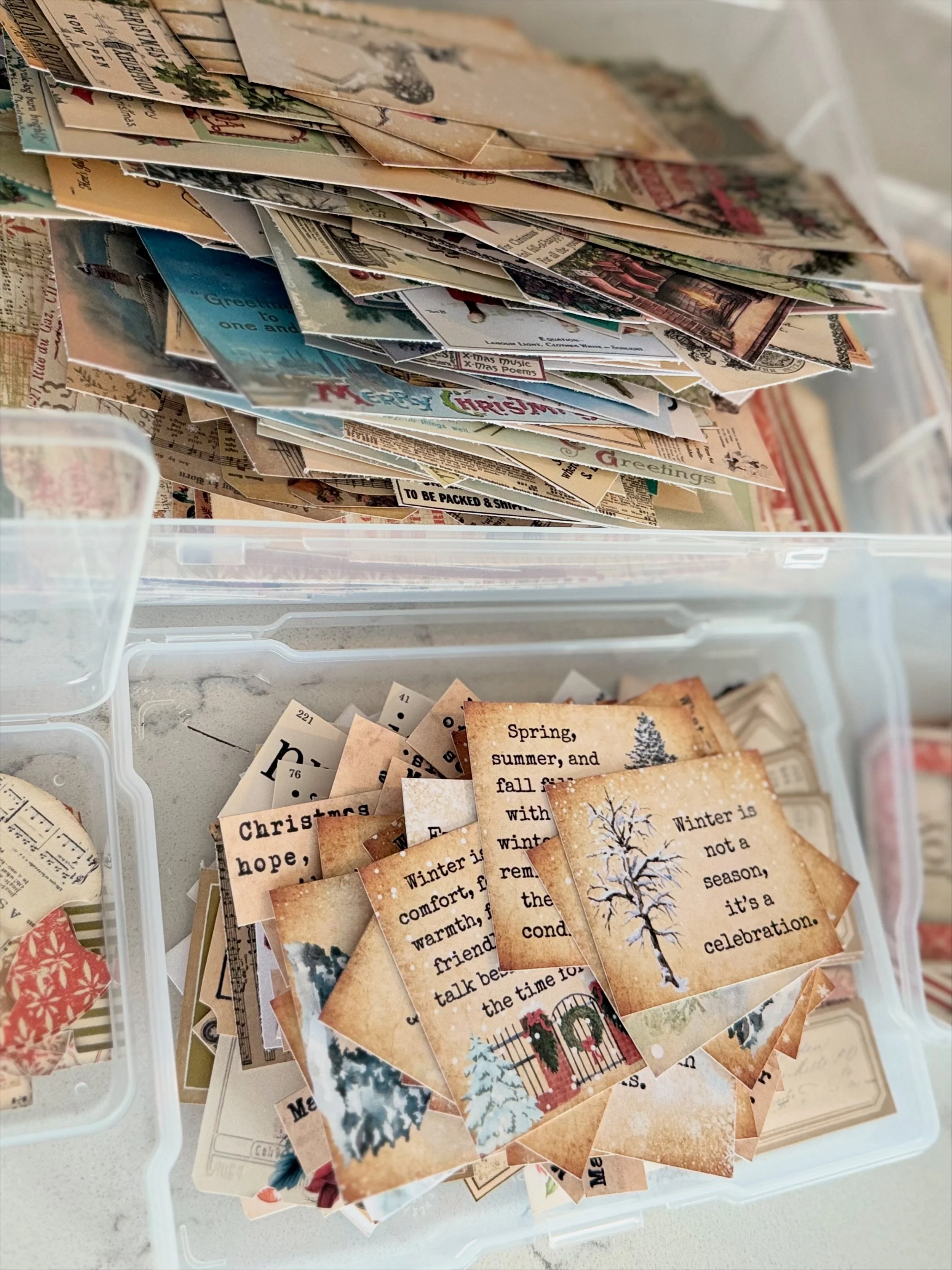 Highlighting holiday journal development. Showcasing plastic storage bins filled with vintage paper scraps, greeting card pieces, printed quotes, and holiday-themed ephemera.