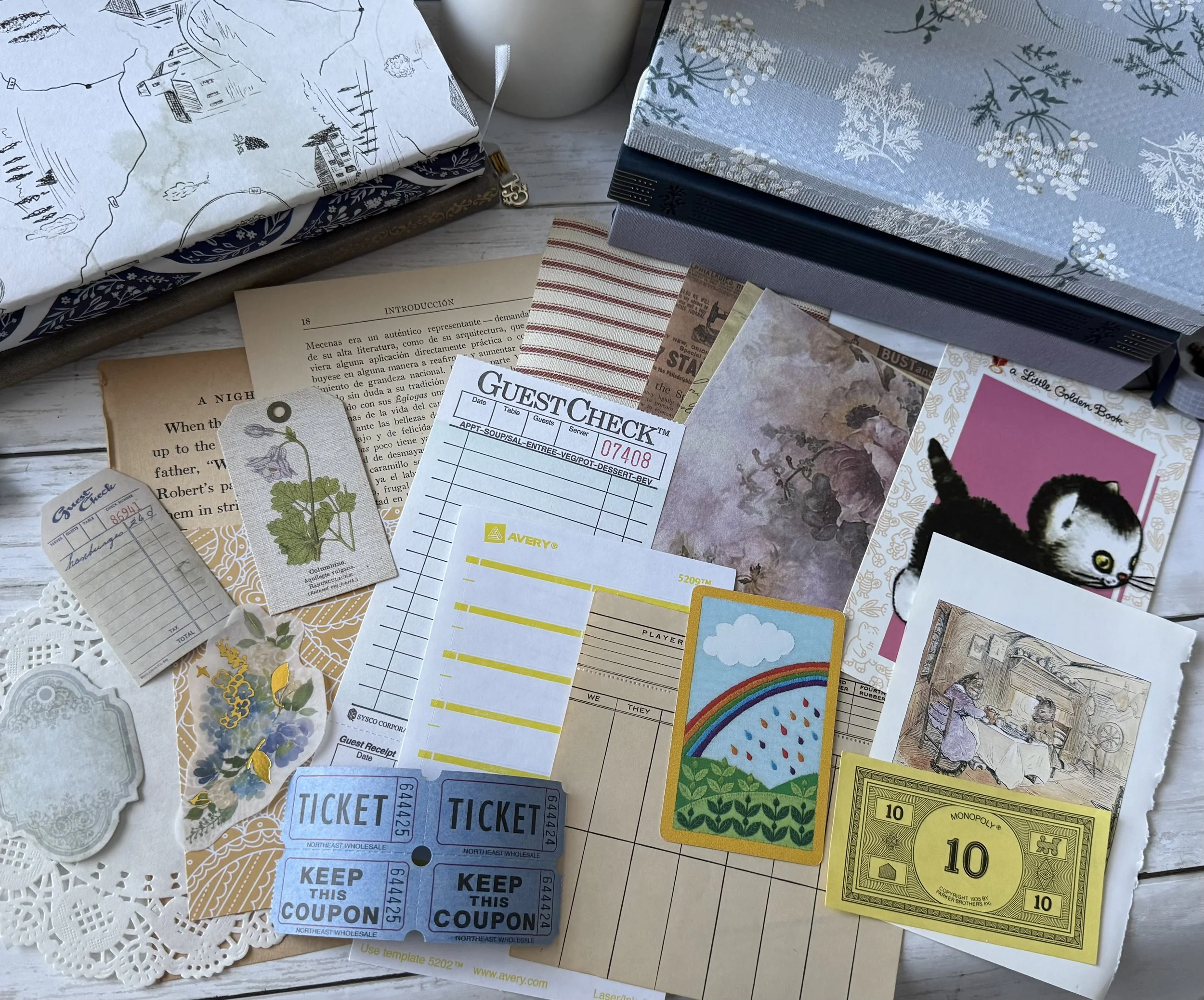 Overhead arrangement of vintage books, floral fabric, restaurant guest checks, tickets, paper ephemera, illustrated cards, and Monopoly money on a white tabletop