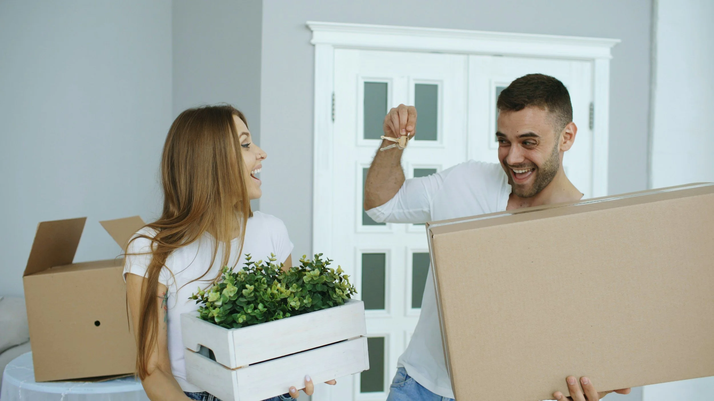 A smiling woman holding a white box with green plants, and a man giving a thumbs up with a large cardboard box, indicating they are moving into a new home.