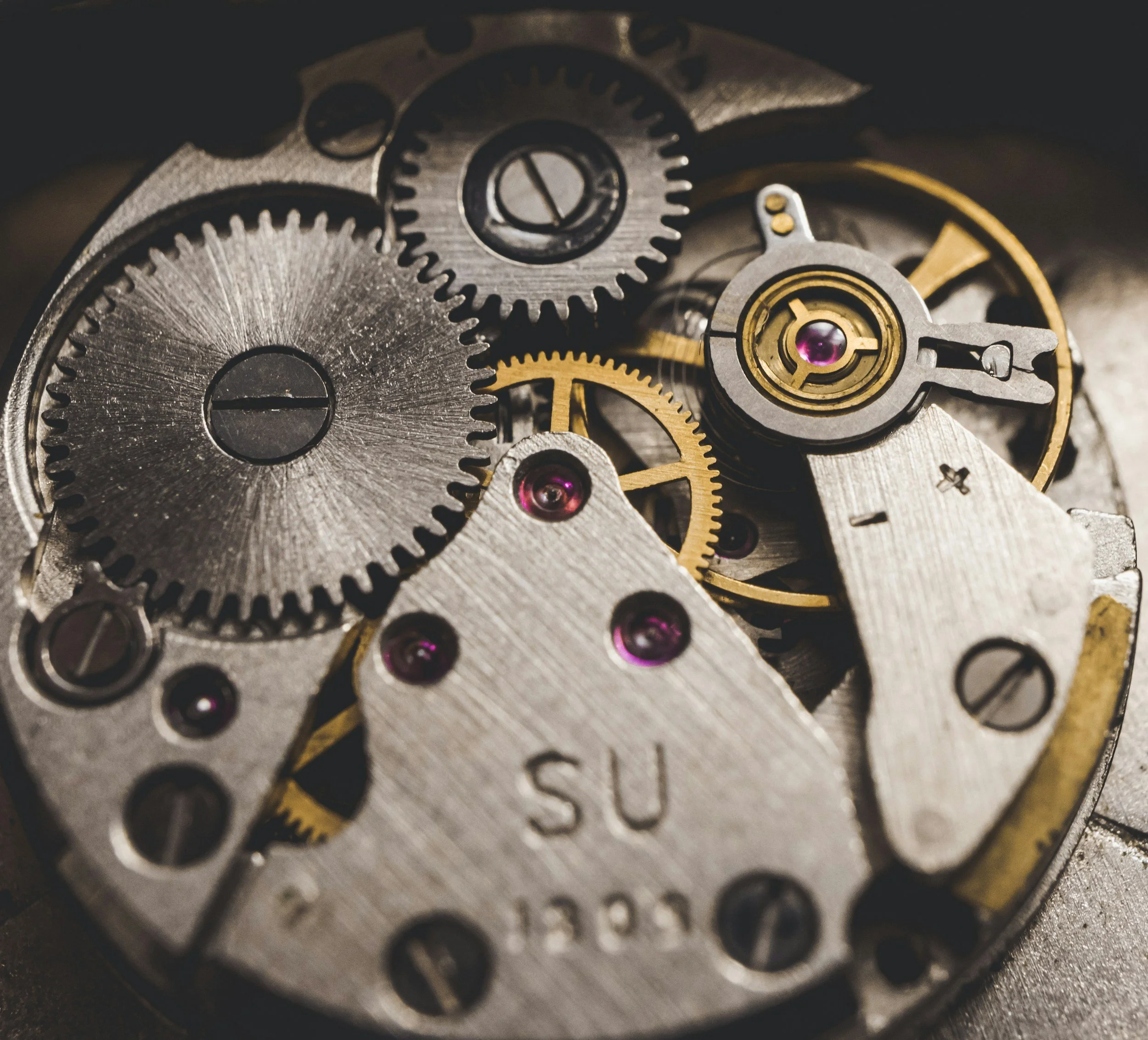 Close-up view of inner gears and mechanical parts inside a watch or clock mechanism, with metallic and gold-colored gears, some with purple jewel bearings.