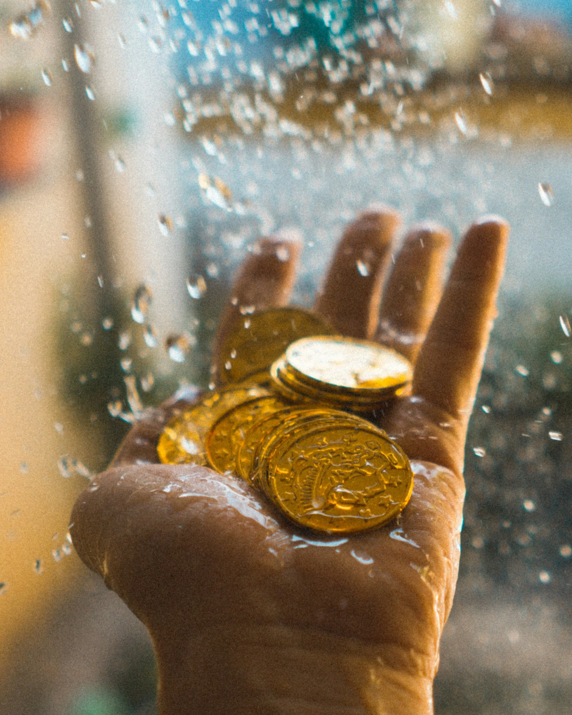 A person's hand holding a handful of gold coins with water droplets on a glass surface in the background.