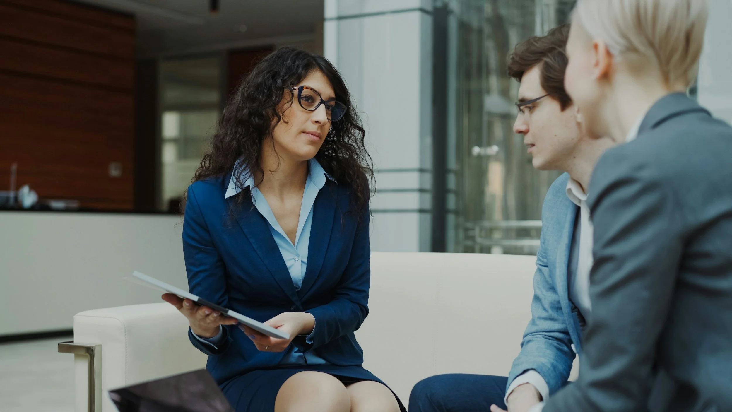 A woman with curly dark hair and glasses dressed in a blue blazer and white shirt sitting on a white chair, holding a tablet, engaged in conversation with two men with glasses, one with light brown hair in a blue sweater and the other with blonde hair in a grey suit jacket, in a modern office setting.