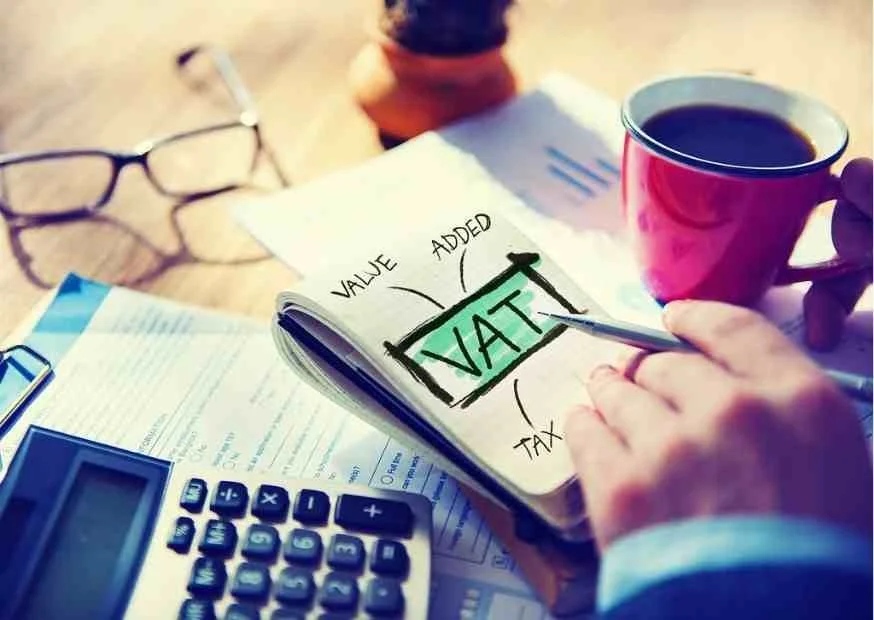 A person filling out a finance or tax document, with a calculator, glasses, and a cup of coffee on a desk.