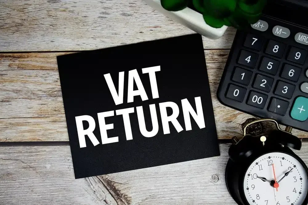 A black paper with white text reading 'VAT RETURN' on a wooden desk. The desk has a calculator, a small clock, and part of a plant in a white pot.