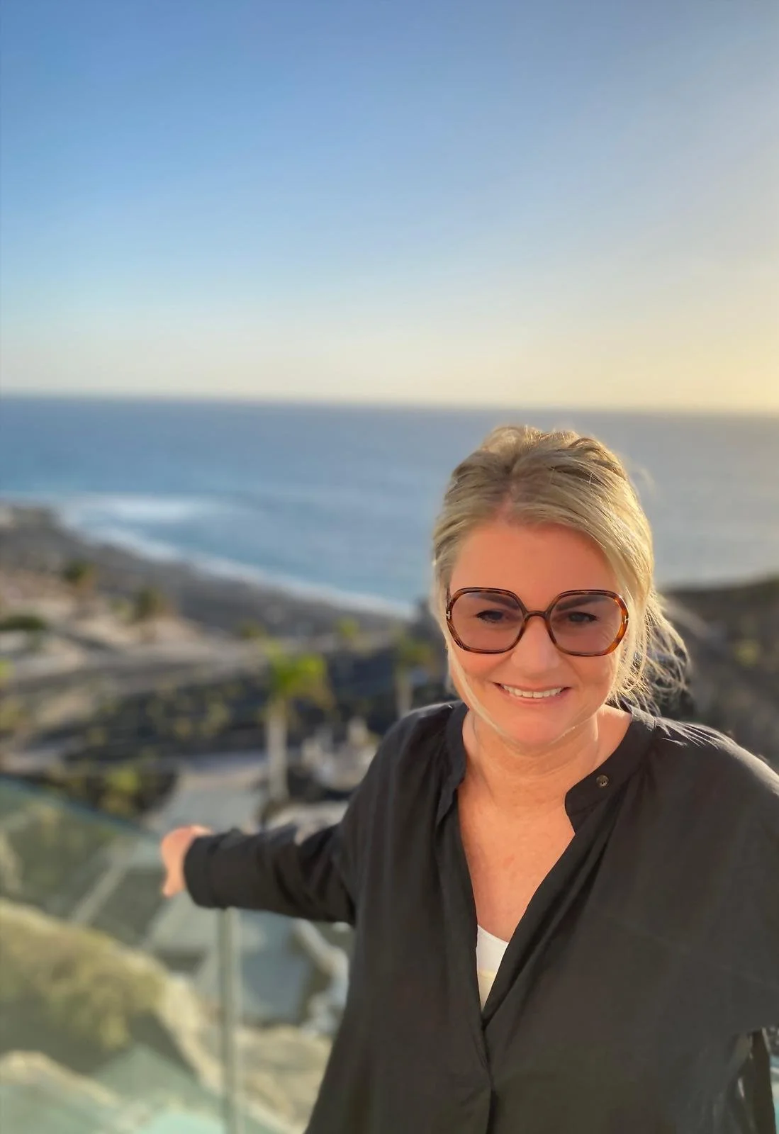 A woman with blonde hair wearing glasses and a black top, smiling at the camera with her arm extended, overlooking a coastal landscape with the ocean, beaches, and a paved path at sunset or sunrise.