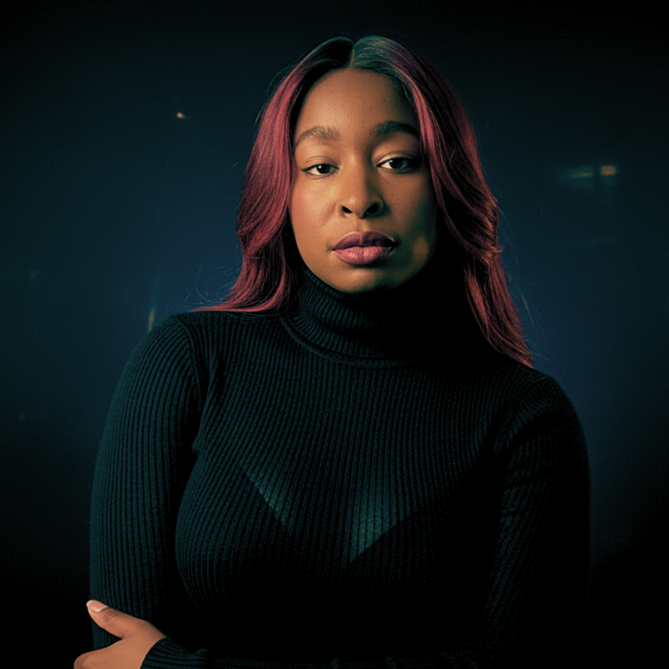 A young woman with long burgundy hair wearing a black turtleneck in a dark studio setting.