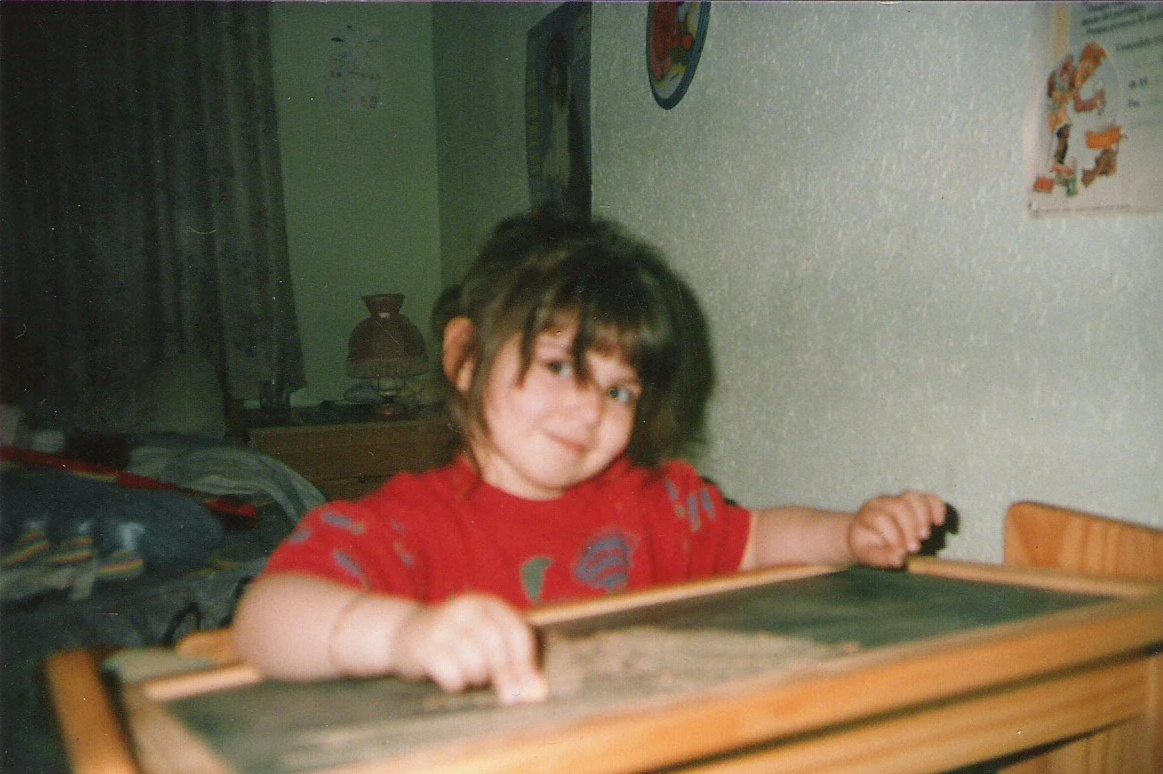 Niña con camiseta roja, con expresión sonriente, sosteniendo una mesa de madera en un ambiente interior.