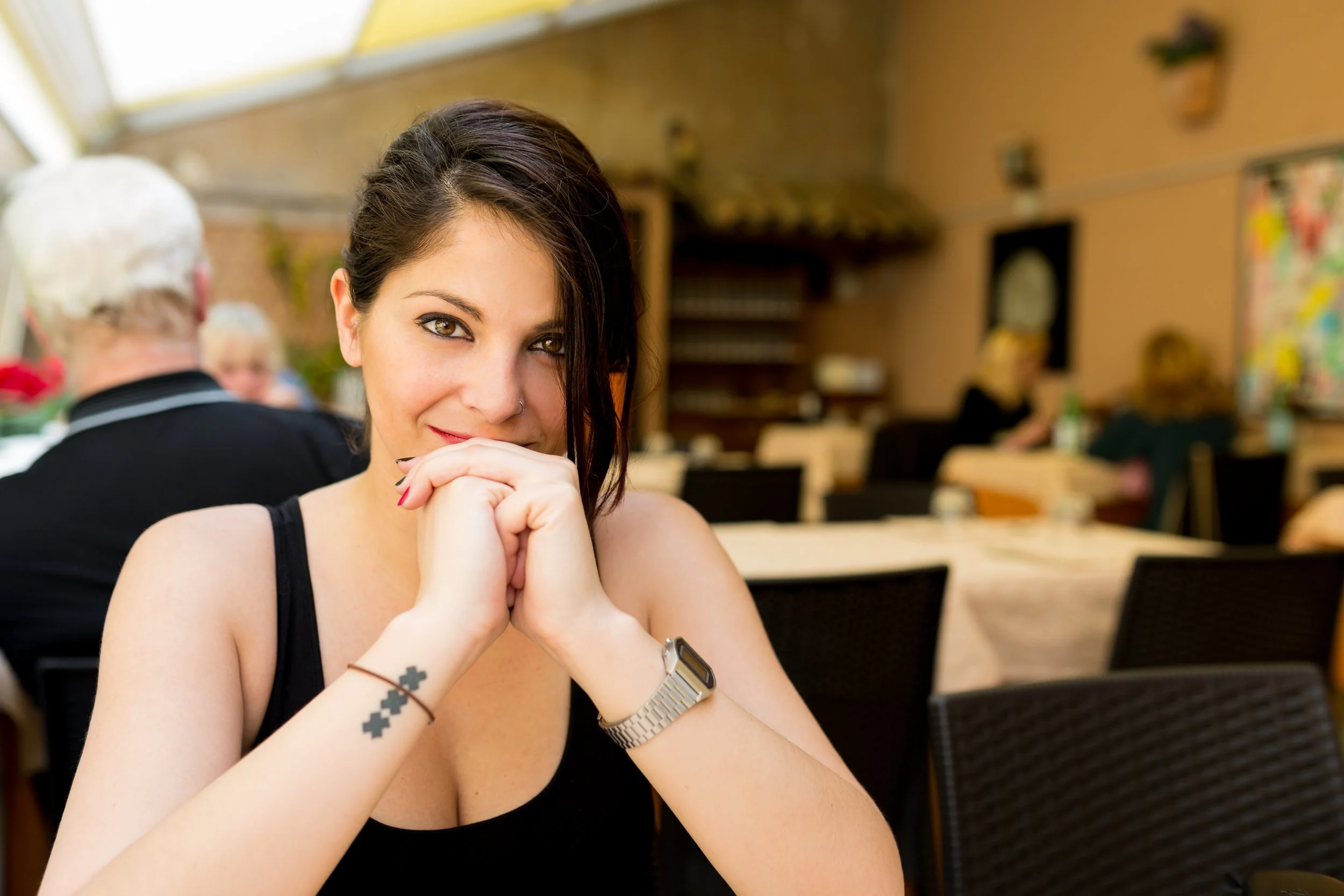 Joven mujer con cabello oscuro, cabello recogido, sin camiseta negra, con tatuaje de cruces en el brazo, y reloj en la muñeca, sonriendo en un restaurante o cafetería con otras personas en el fondo.