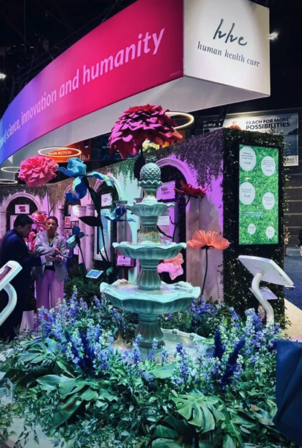 Exhibit at a trade show featuring colorful floral decorations, a multi-tiered fountain, and large pink signs with messages about humanity and health care.