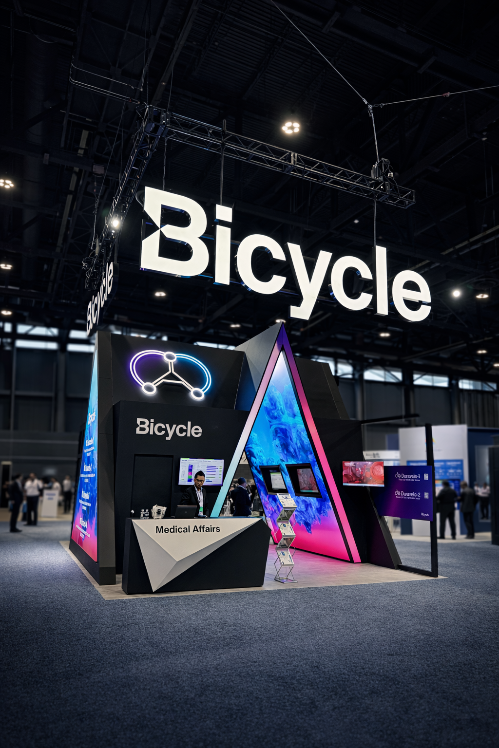 A trade show booth with the prominent word 'Bicycle' illuminated on a large hanging sign and on the booth front. The booth features digital screens, neon lights, and a sign that says 'Medical Affairs,' with a person working at the desk. The background shows a busy exhibition hall.