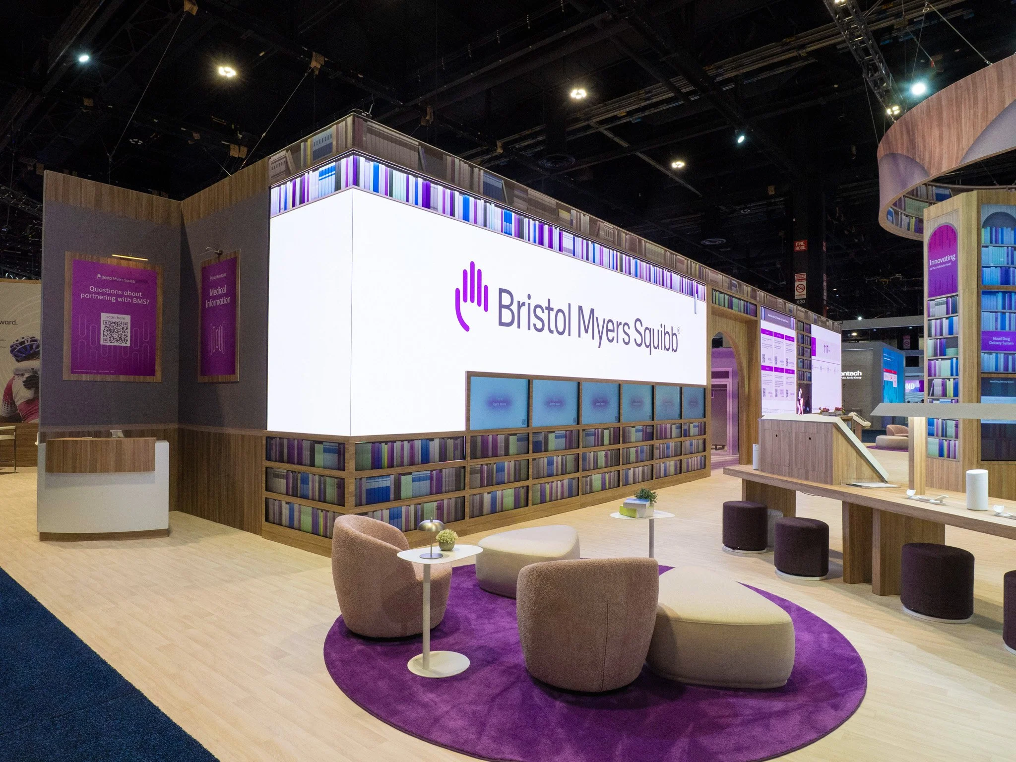 Exhibition booth for Bristol Myers Squibb featuring a large digital screen with the company's logo, bookshelves, lounge area with chairs and tables, and informational displays.