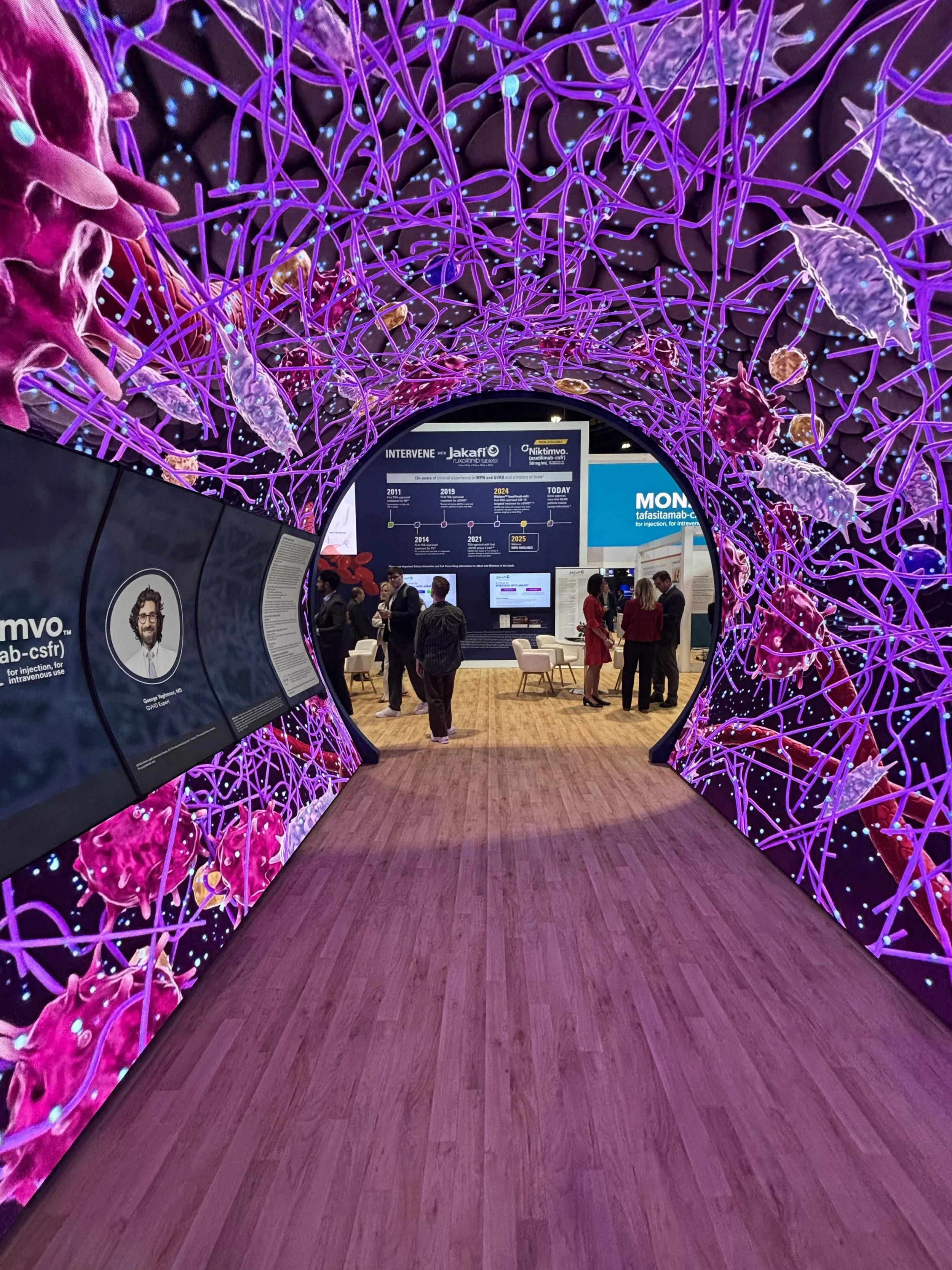 A futuristic tunnel exhibit with bright purple and pink neural network-like graphics overhead, leading to a conference or expo with people discussing and informational displays.