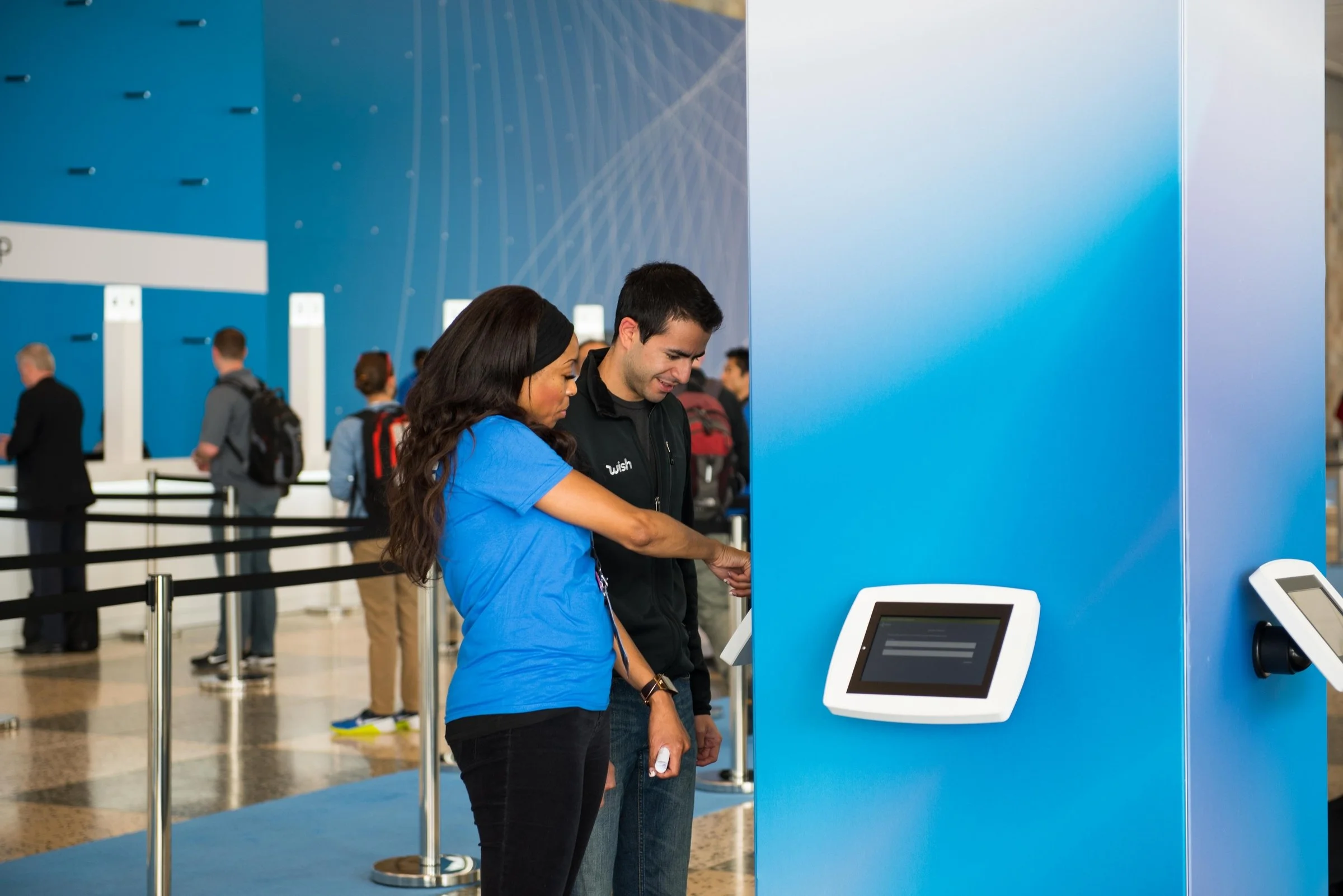 Two people using a self check-in kiosk at an airport, with other travelers and check-in counters in the background.
