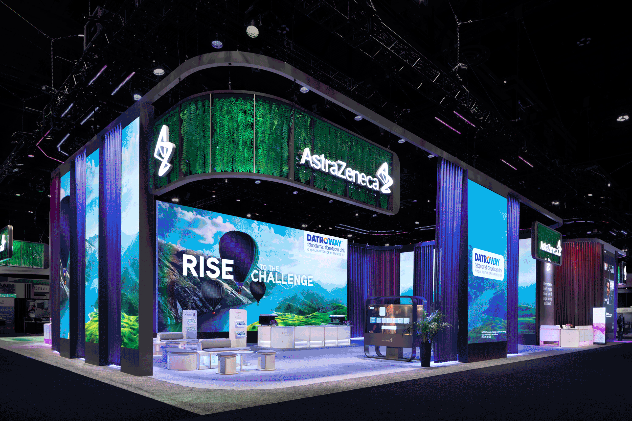 Trade show booth for AstraZeneca with digital screens displaying scenic landscapes and the slogan "Rise to the challenge" with hot air balloons, set in a large exhibition hall.
