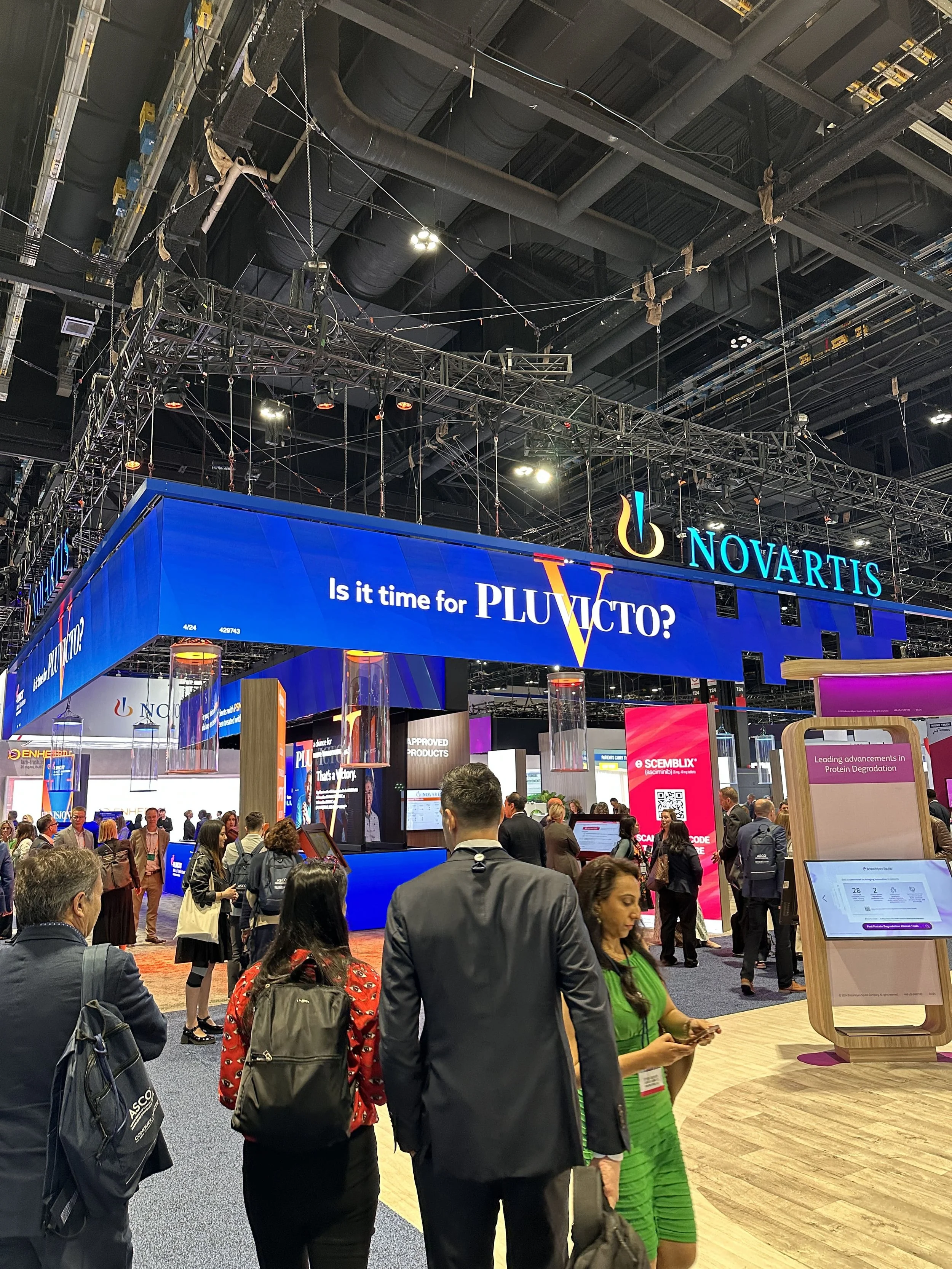 A busy trade show with a large blue booth for Novartis, featuring the text 'Is it time for PLUVTO?'. There are people walking around, some engaging with displays and others talking. The environment has high ceilings with industrial lighting and rigging.