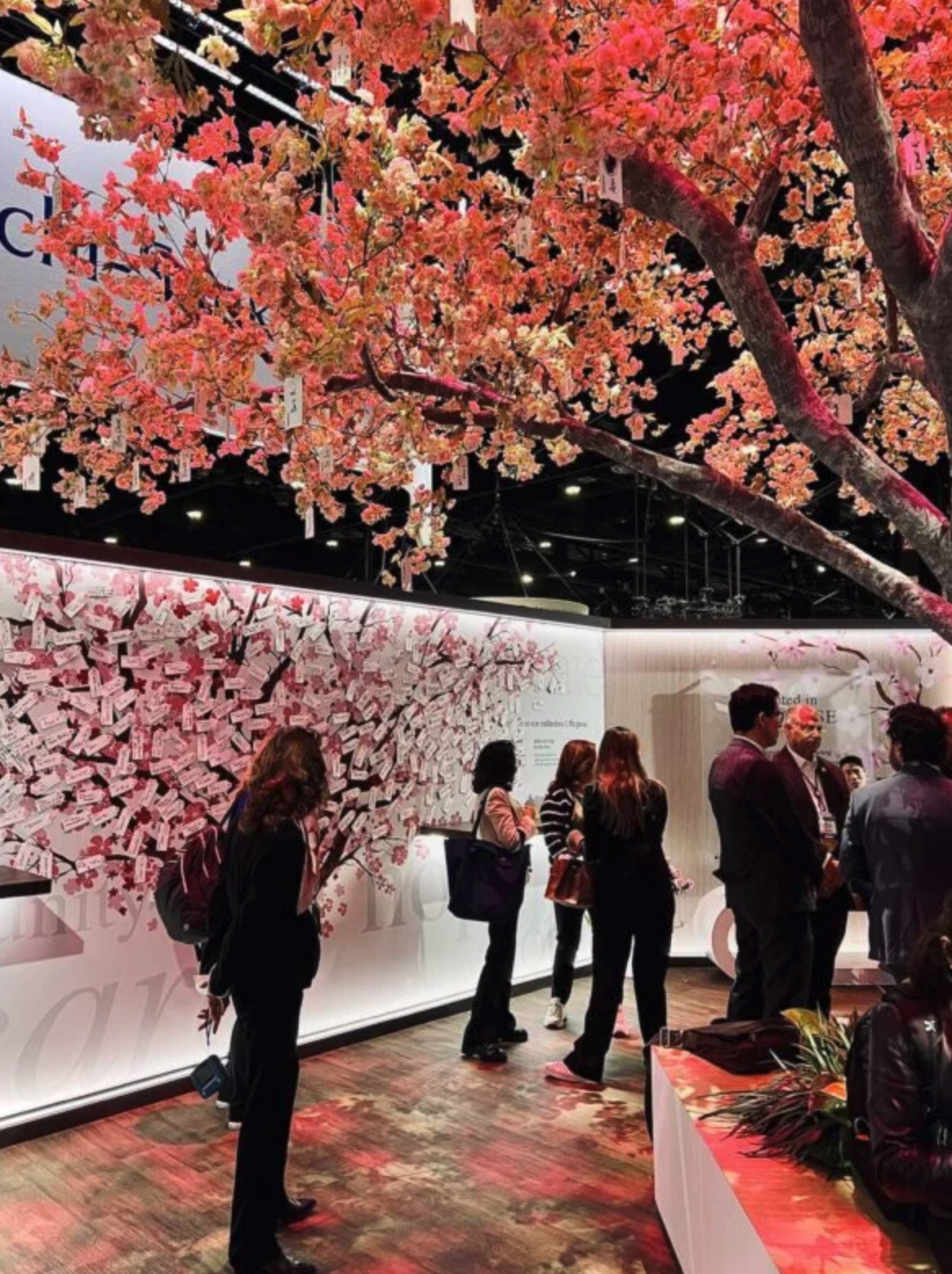 People at a display booth with pink cherry blossom tree and wall installation decorated with pink and white paper notes or cards, at an indoor event or exhibition.
