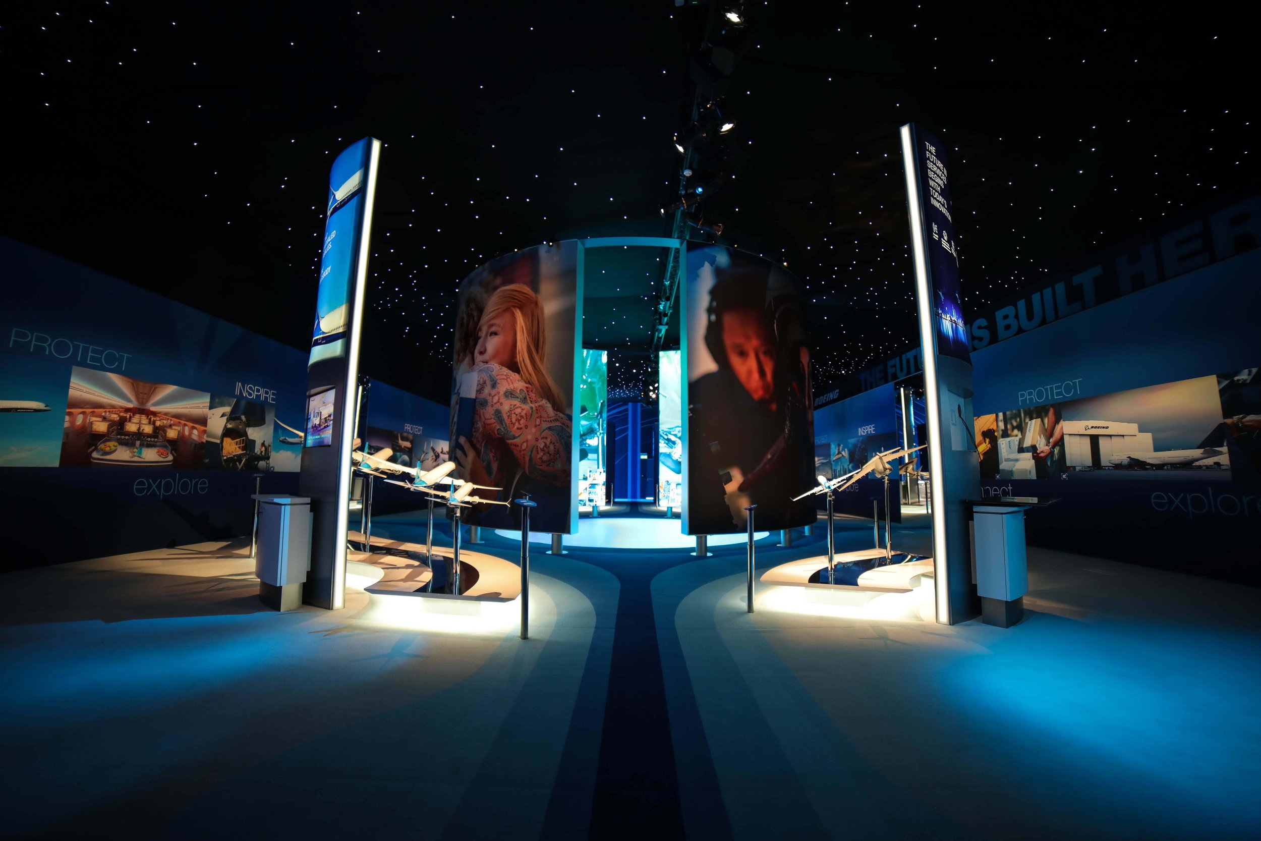 An indoor exhibition space featuring large curved digital screens displaying photos of people, with lighting and display panels showcasing aerospace and technology themes, and a starry ceiling overhead.