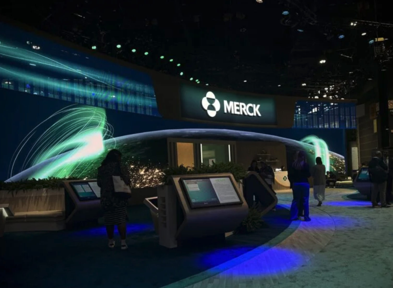 Merck exhibition booth at a conference with digital displays, people viewing screens, and the Merck logo prominently displayed.