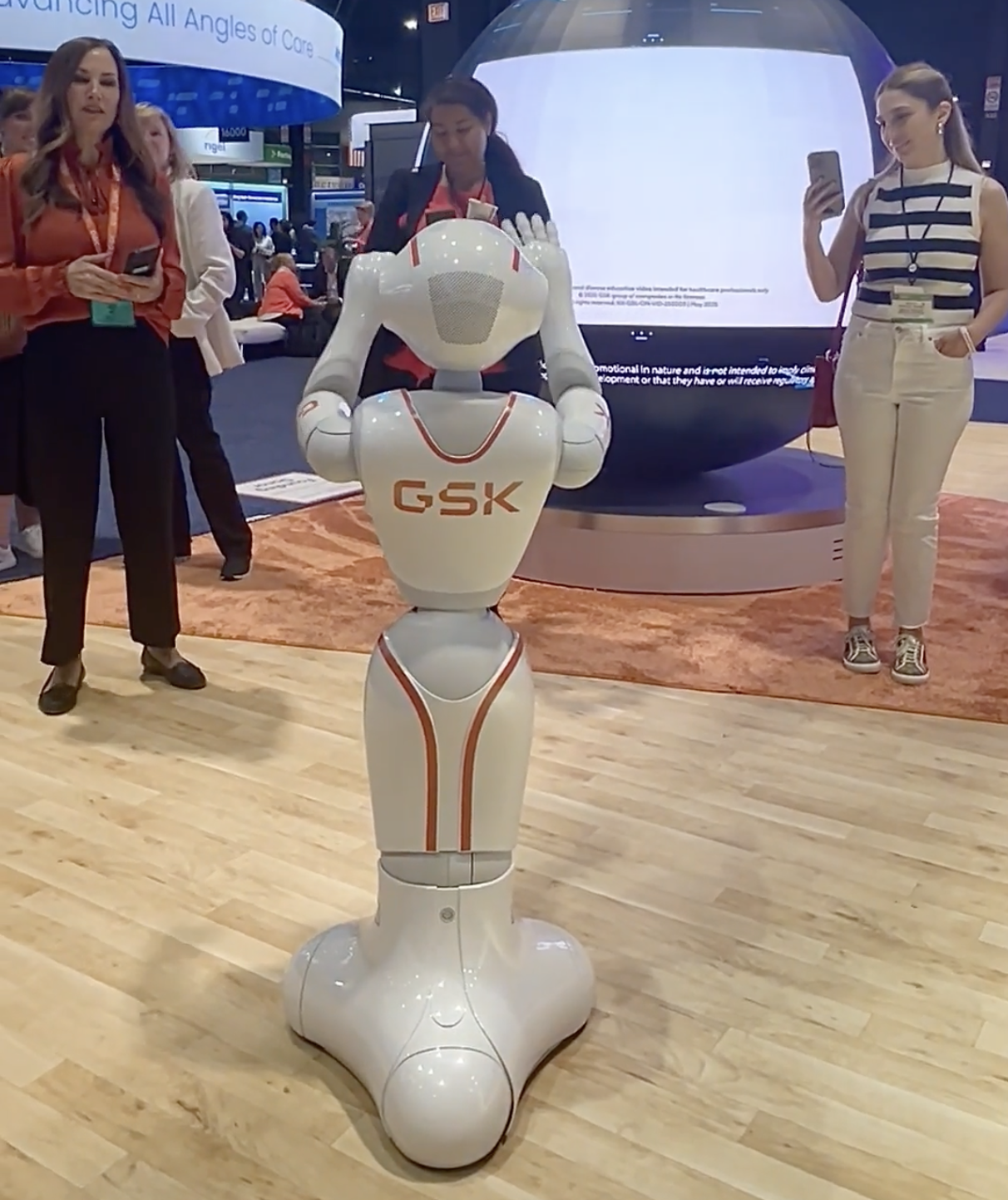 A robot with the logo GSK standing on a wooden floor at a conference or event, with two women nearby taking photos and looking at their phones.