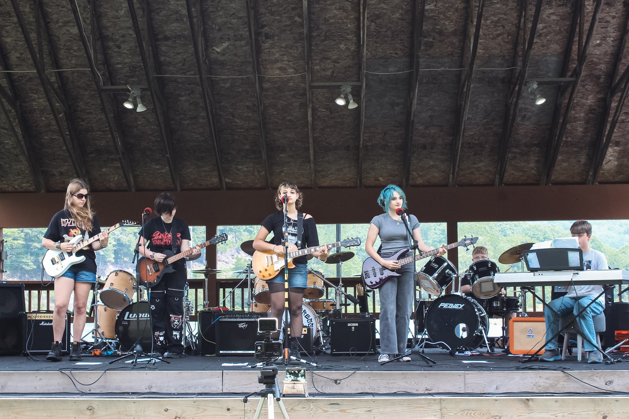A band of six musicians performing on an outdoor stage, with three women playing guitars and drums and two men on keyboard and drums, under a wooden roof with trees visible in the background.