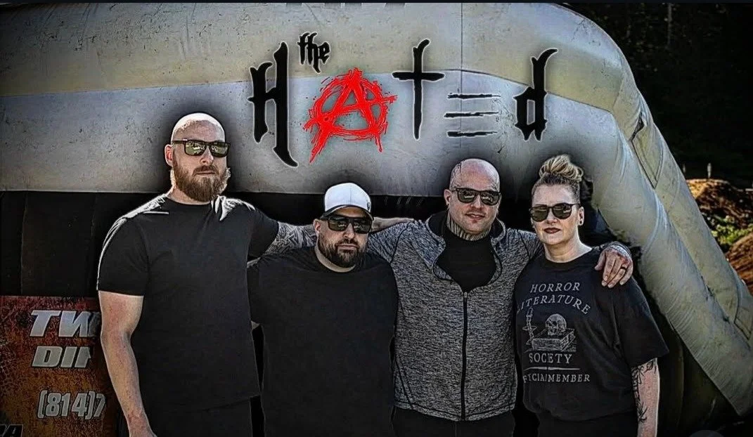 Four people standing in front of a trailer with 'the floated' written on it. They are wearing dark clothing and sunglasses, with one person wearing a shirt that says 'Horror Literature Society'.