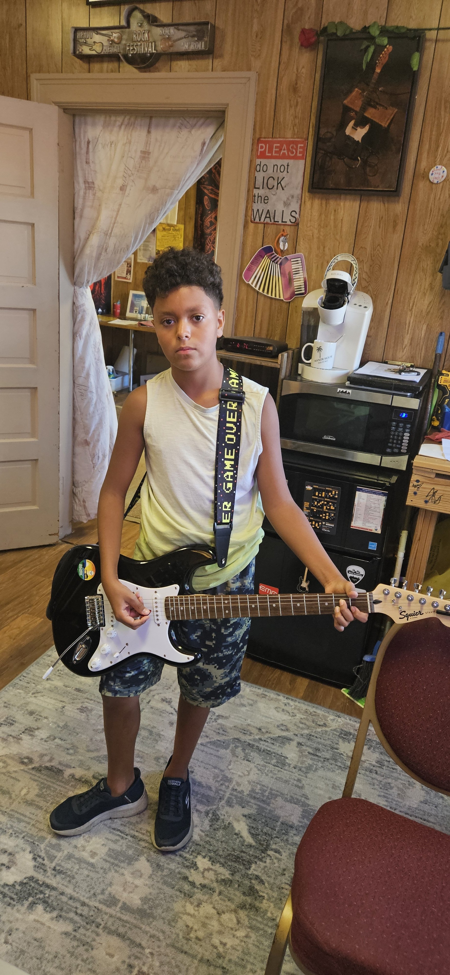 A young boy standing indoors holding a black and white electric guitar, wearing a sleeveless shirt, camouflage shorts, and black sneakers, in a room with wooden panel walls decorated with music-themed signs and artwork, a microwave, and a sign that s