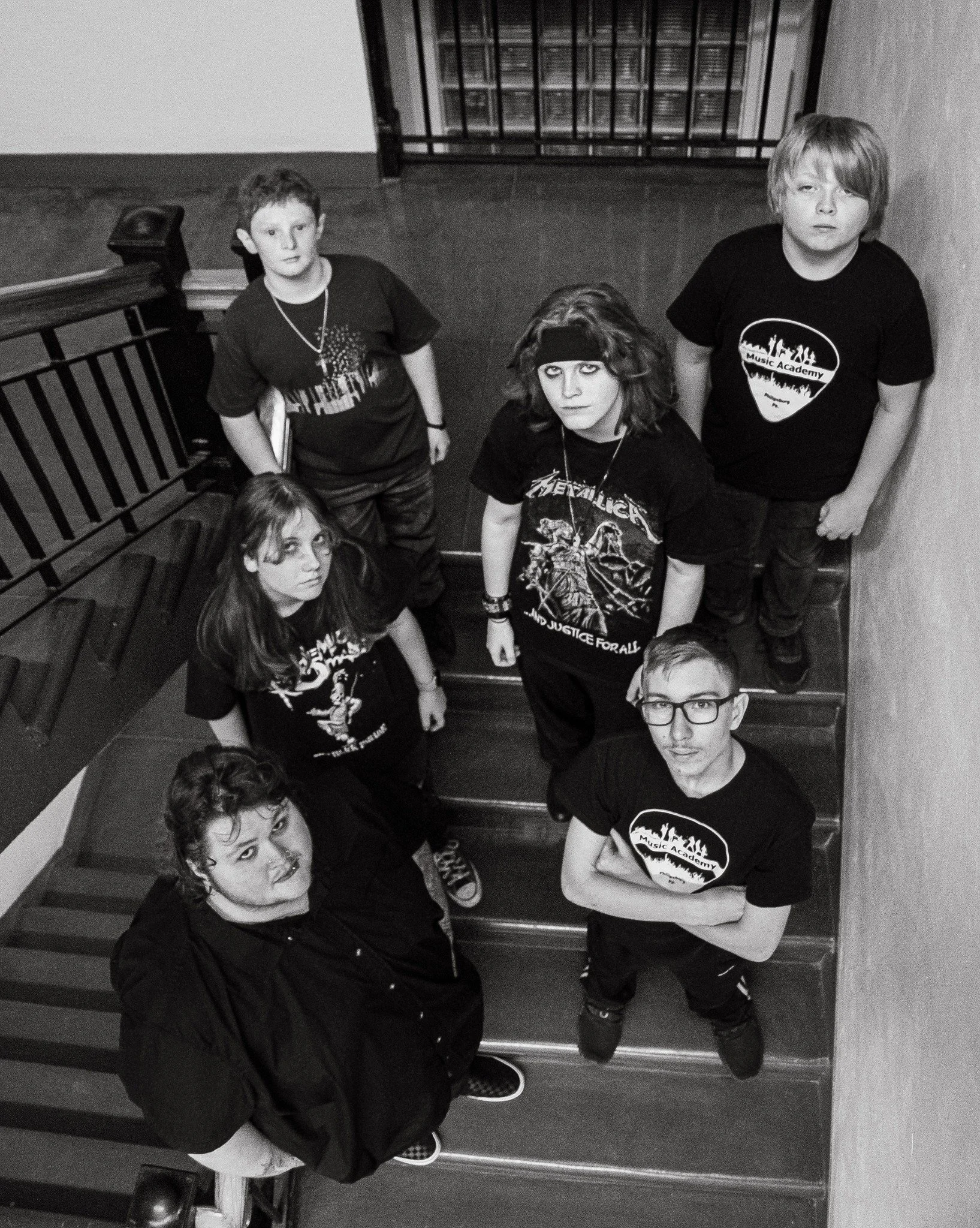 Group of six young people standing on a staircase, looking up at the camera. They are wearing dark clothing with band logos and accessories, with a grunge or punk style.