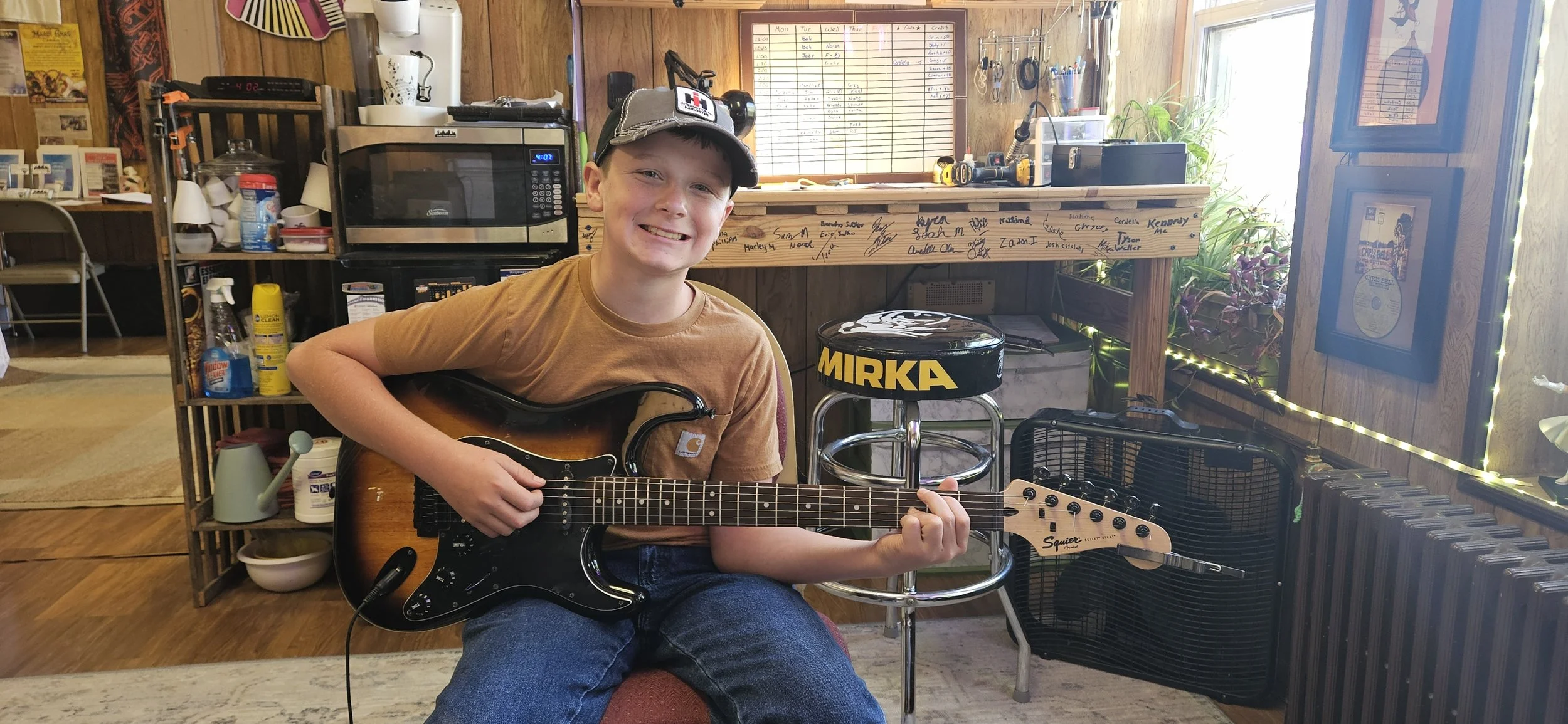 Young boy sitting on a stool playing an electric guitar in a cozy, cluttered room with wood-paneled walls, a microwave, a whiteboard, and various music equipment and decorations.