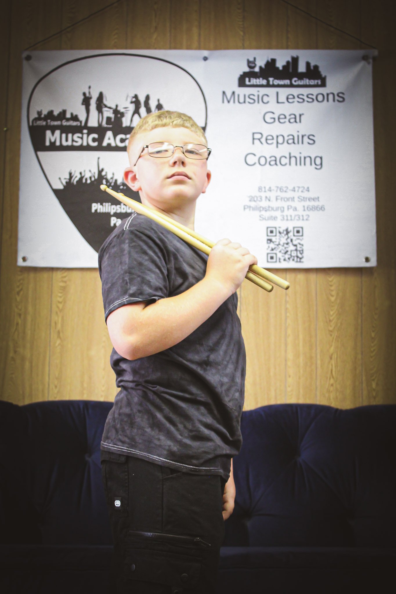 A young boy wearing glasses and a black t-shirt holding a yellow drumstick in his right hand. He is standing in front of a wooden wall with a sign that advertises music lessons, gear, repairs, and coaching at Little Town Guitars in Philipsburg, PA.
