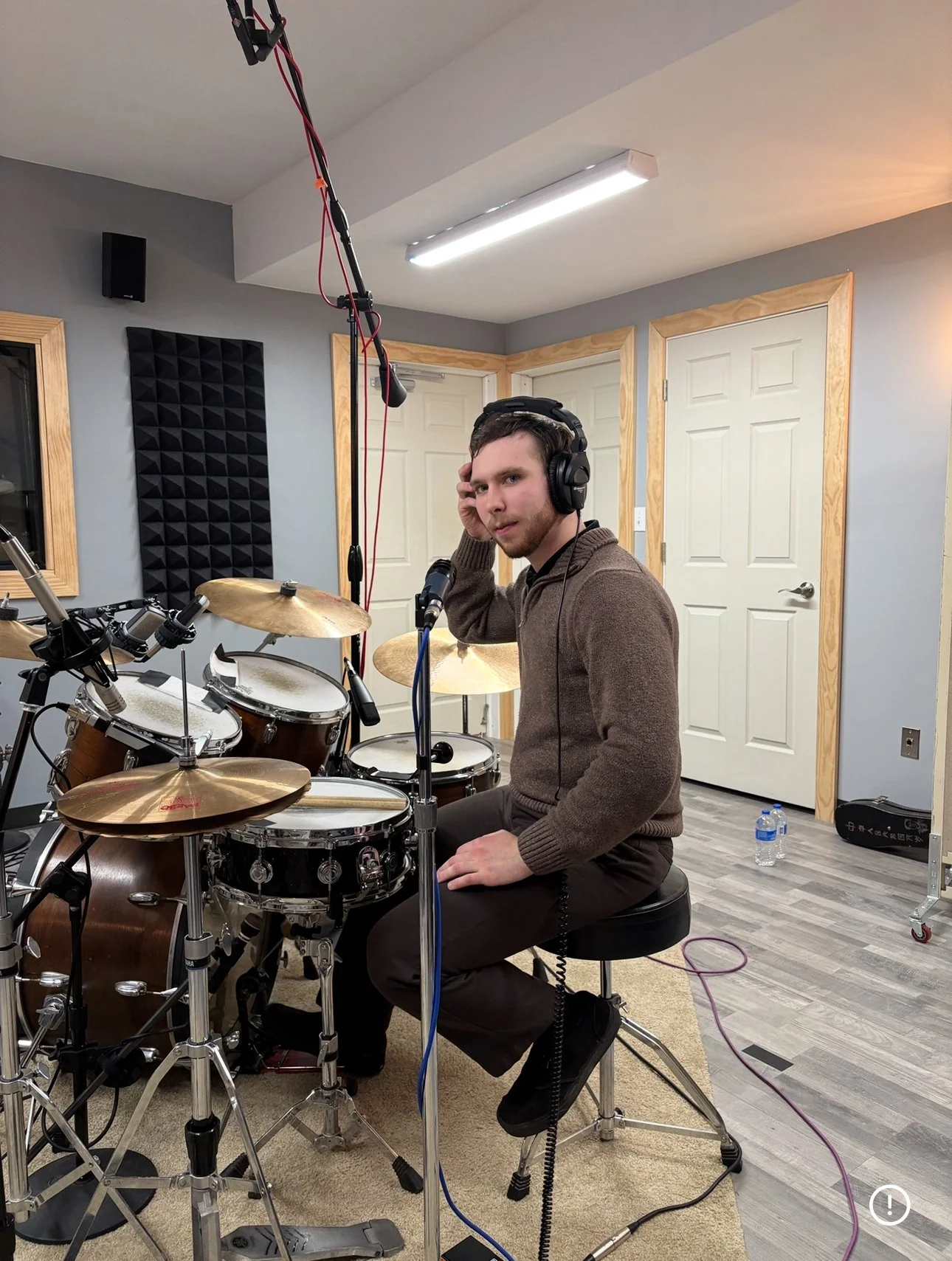 A man sitting behind a drum set in a recording studio, wearing headphones and adjusting one ear of the headphones, with microphones and recording equipment around him.