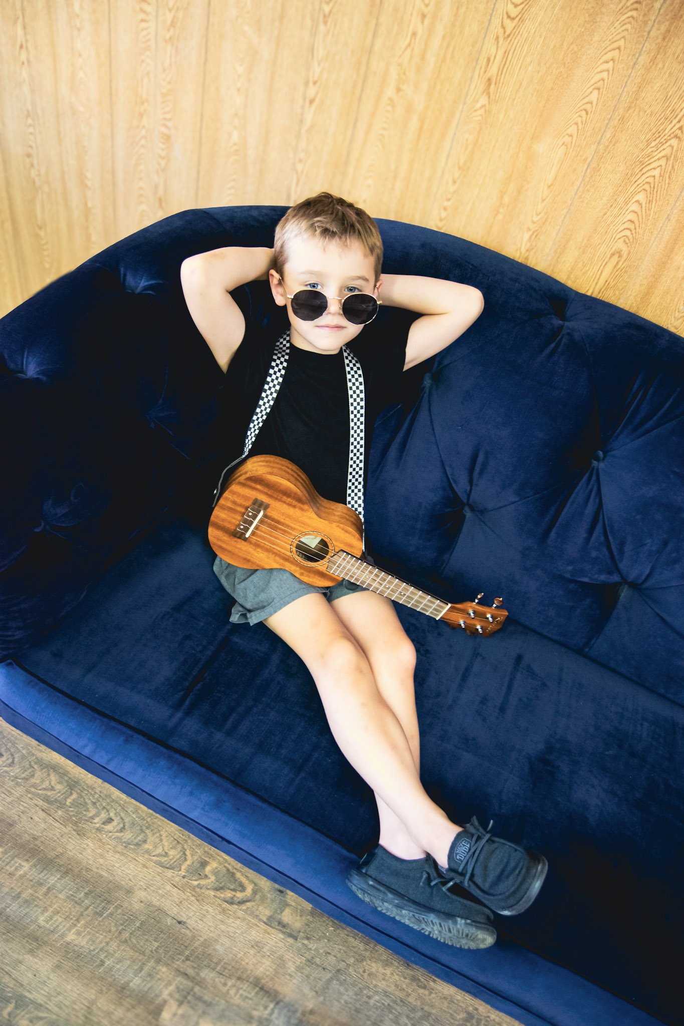 A young boy lying on a navy blue couch with his hands behind his head, wearing sunglasses and black t-shirt, holding a small wooden ukulele.