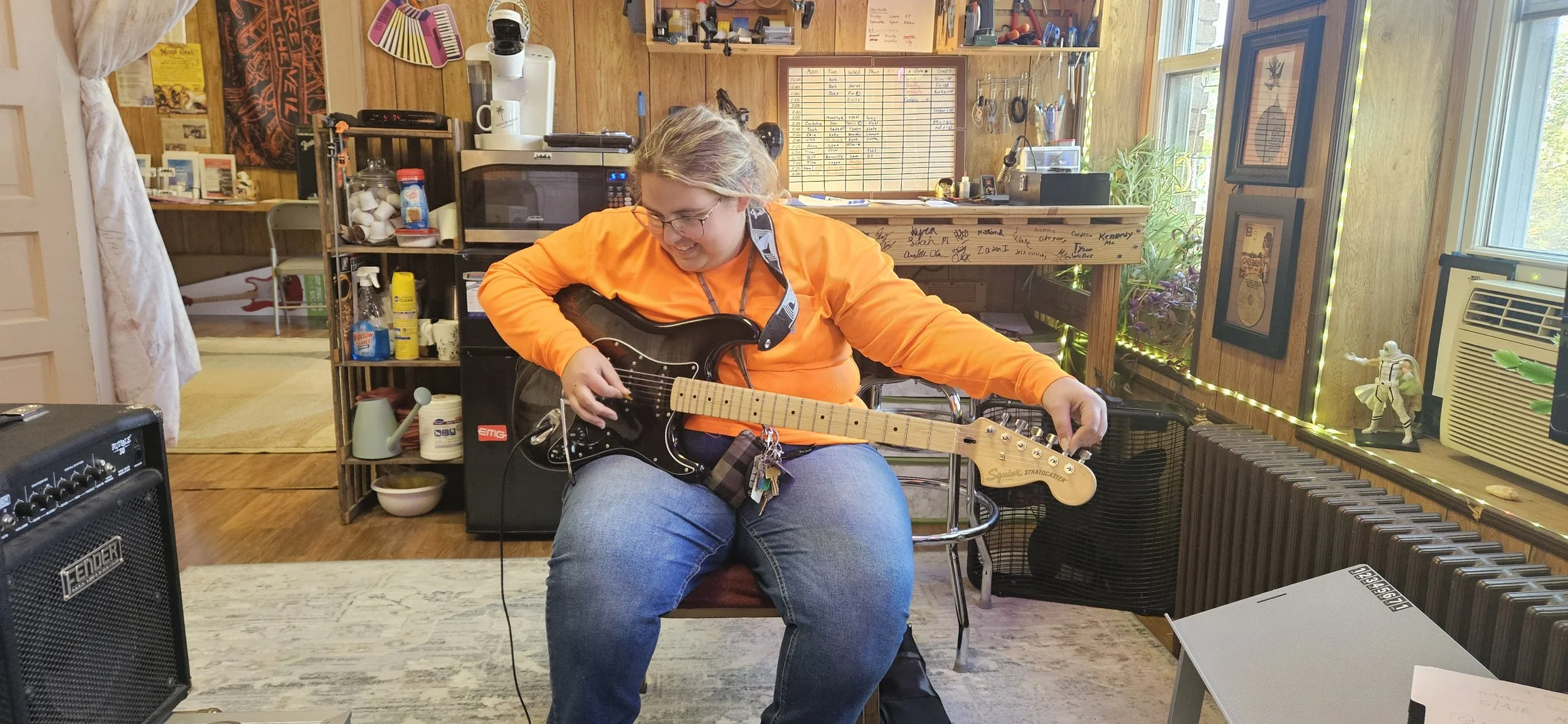 A woman sitting on a chair in a cozy, wood-paneled room, playing a black electric guitar. She is wearing an orange long-sleeve shirt and glasses, with a keychain hanging from her belt. The room has various decorations, shelves with items, a window wi