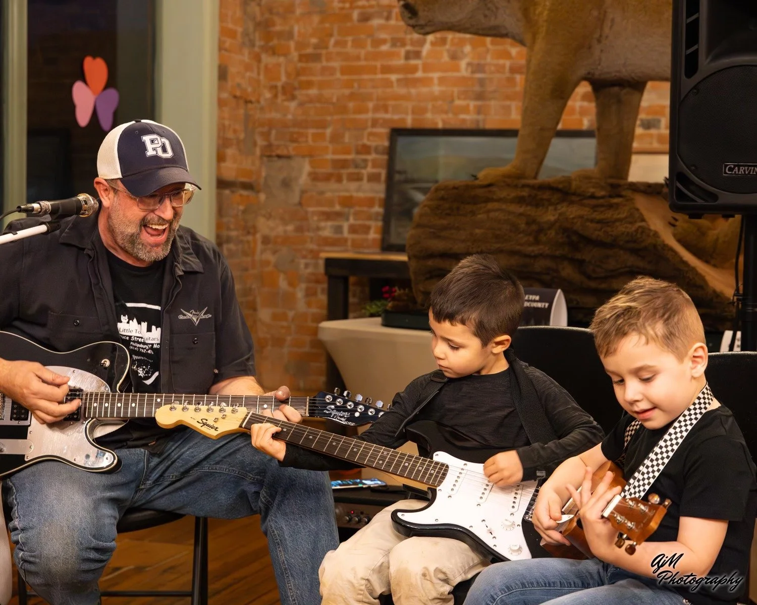 A man with glasses, wearing a baseball cap and black shirt, playing an electric guitar while singing, with two young boys sitting next to him, also playing electric guitars, in a room with brick walls, a large rock formation, and a speaker.