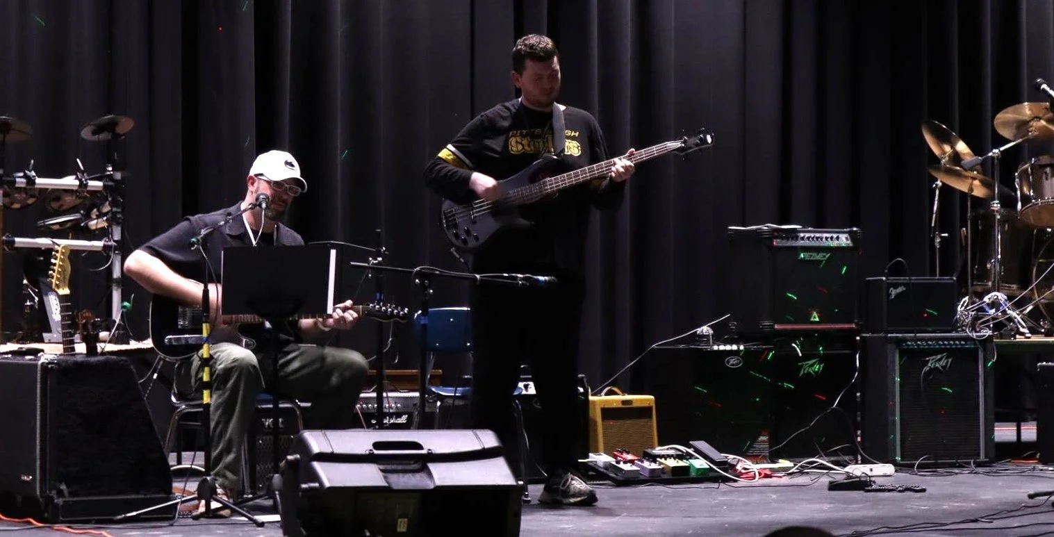 Two musicians performing on stage with various electronic equipment and drums, one playing acoustic guitar and the other playing bass guitar.