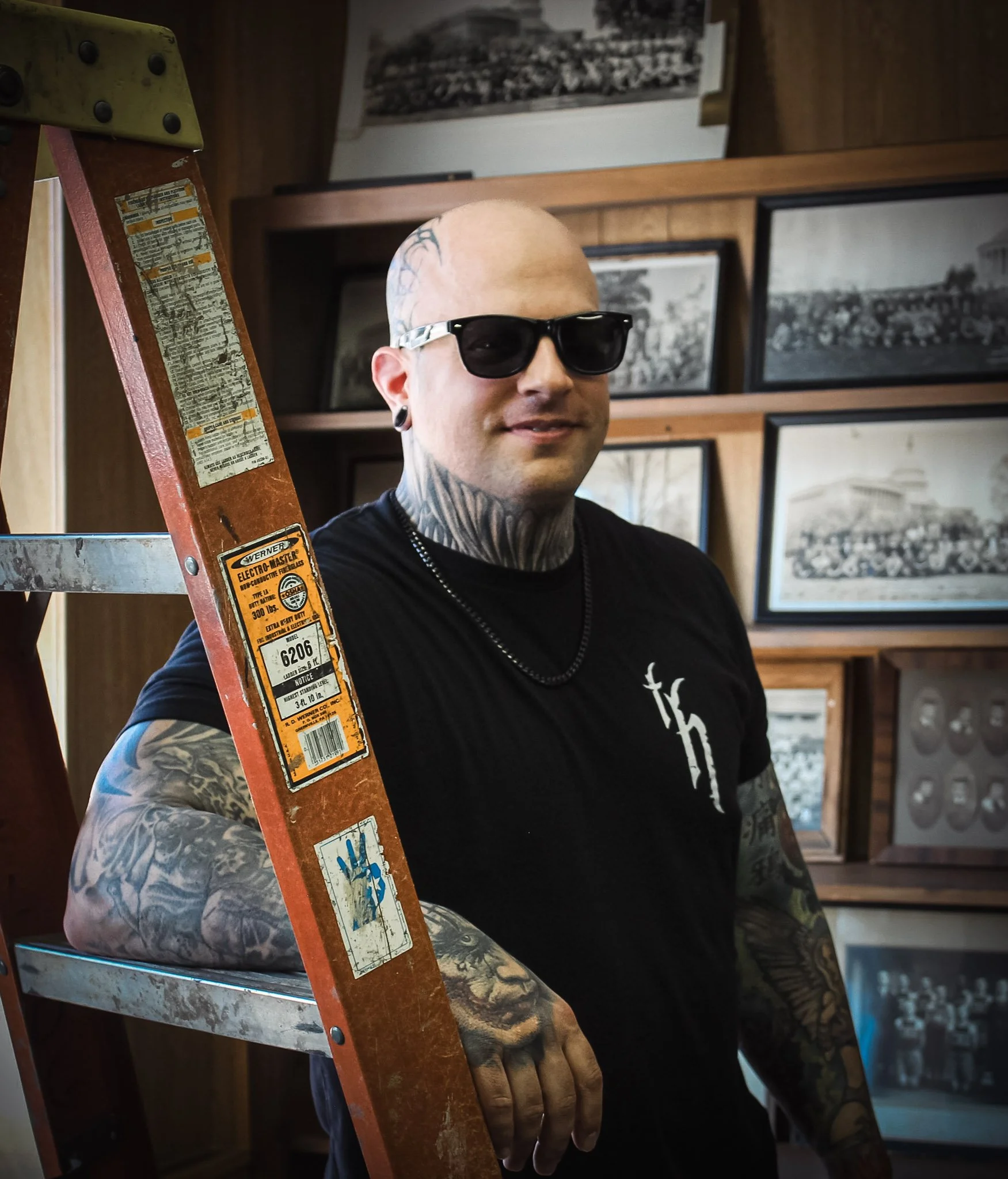 Man with tattoos and sunglasses standing next to a ladder indoors with framed photos on the wall.
