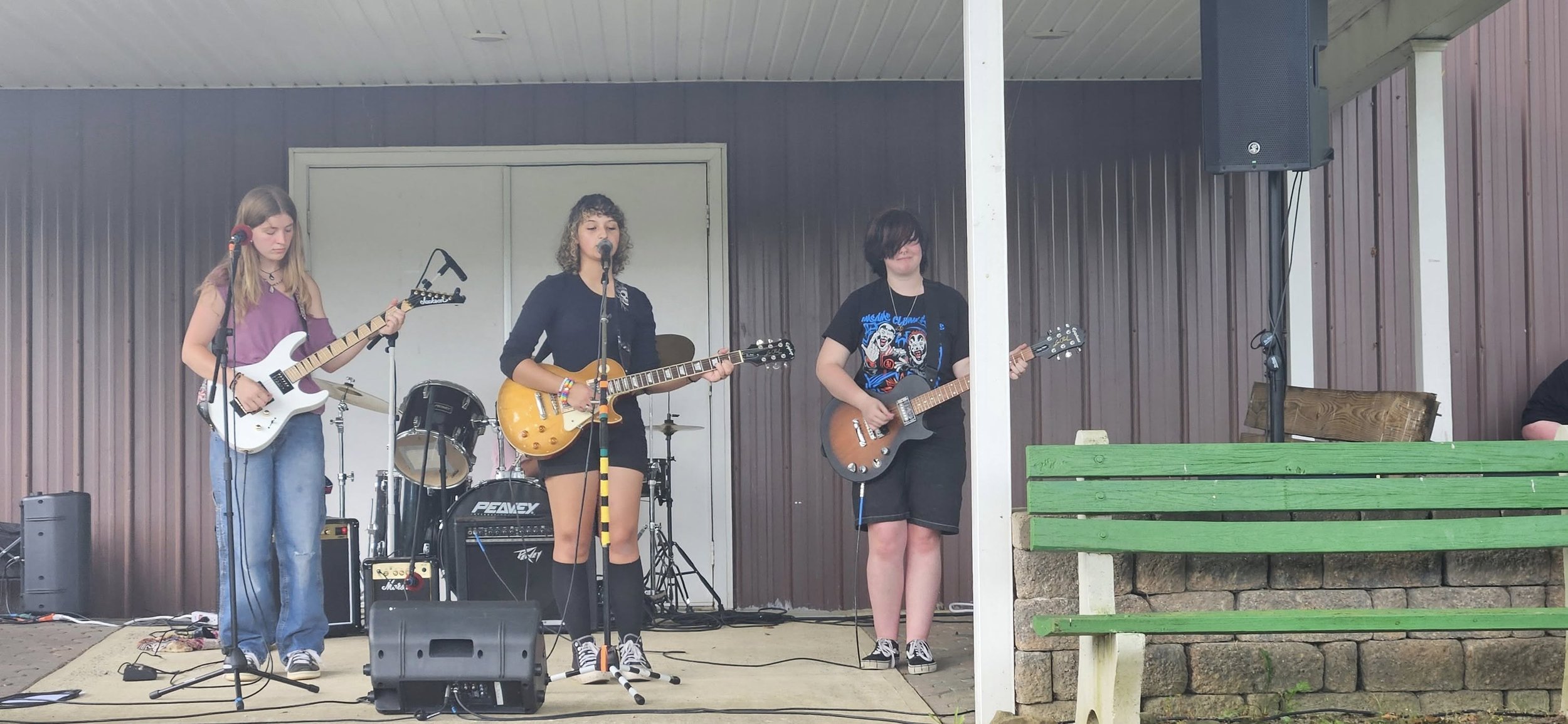 A group of four young people performing music on stage with guitars and drums, outdoors under a roofed stage area with brown and purple siding and a green bench in front.