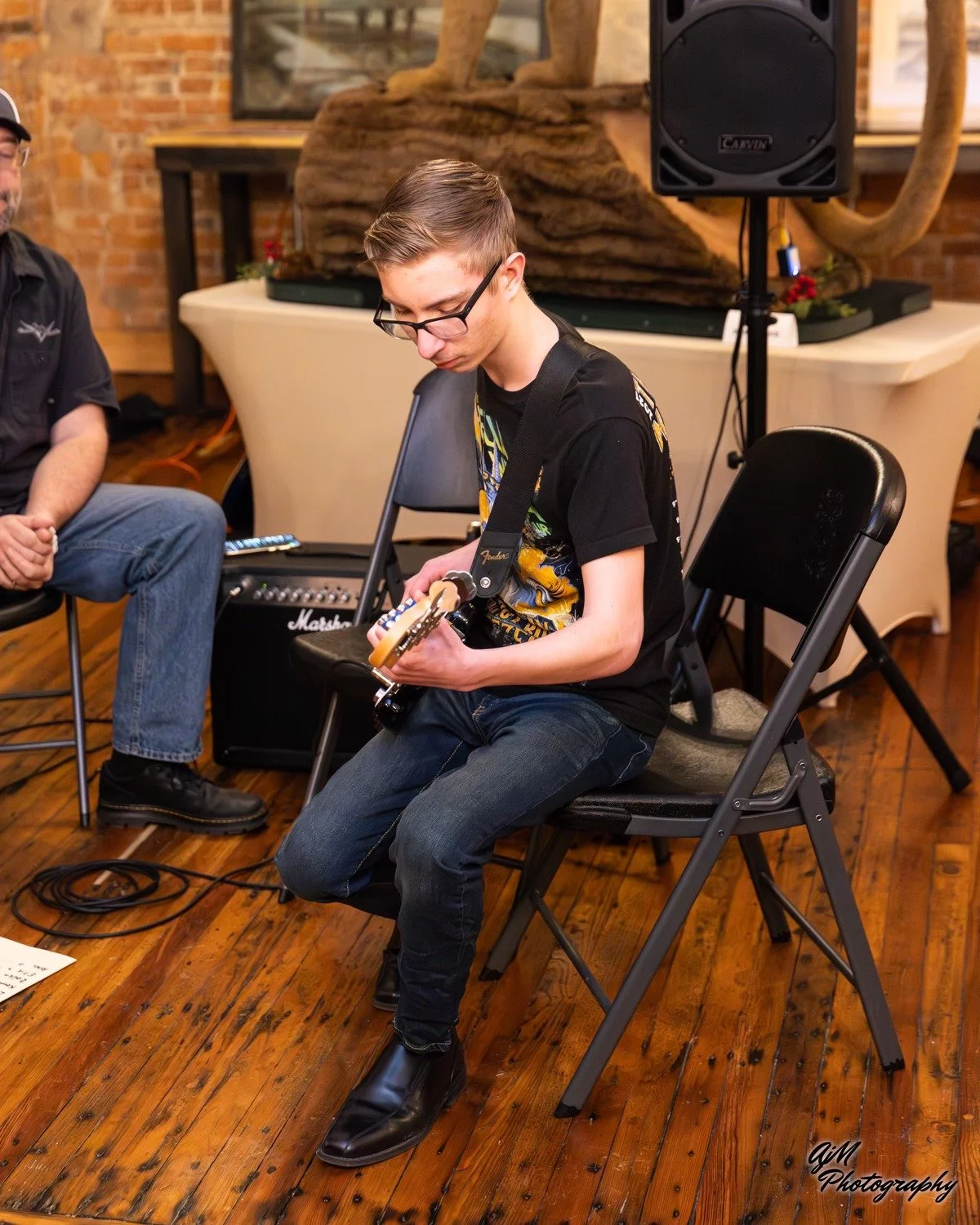 A young man wearing glasses and a black T-shirt playing an electric guitar while sitting on a black folding chair in a room with wooden floors and brick walls.