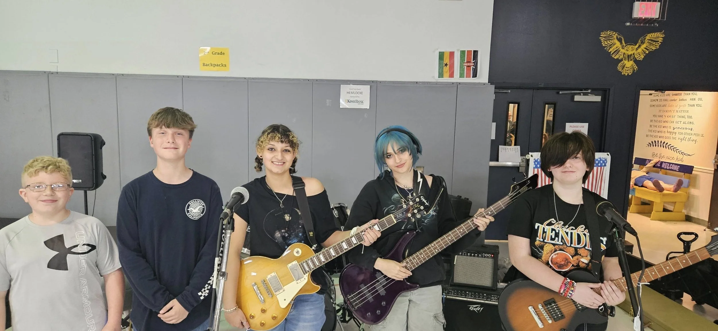 Four young people posing with musical instruments in a school gymnasium, two holding guitars and one holding a bass guitar, all smiling, with a doorway in the background.