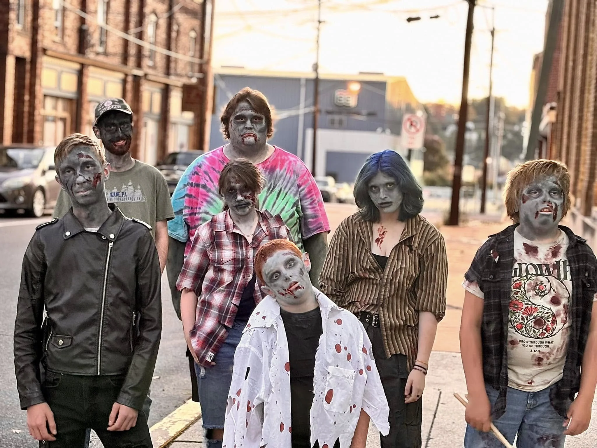 A group of eight kids dressed up as zombies, with makeup resembling rotting flesh and blood, standing outdoors on a street at sunset, with buildings and cars in the background.