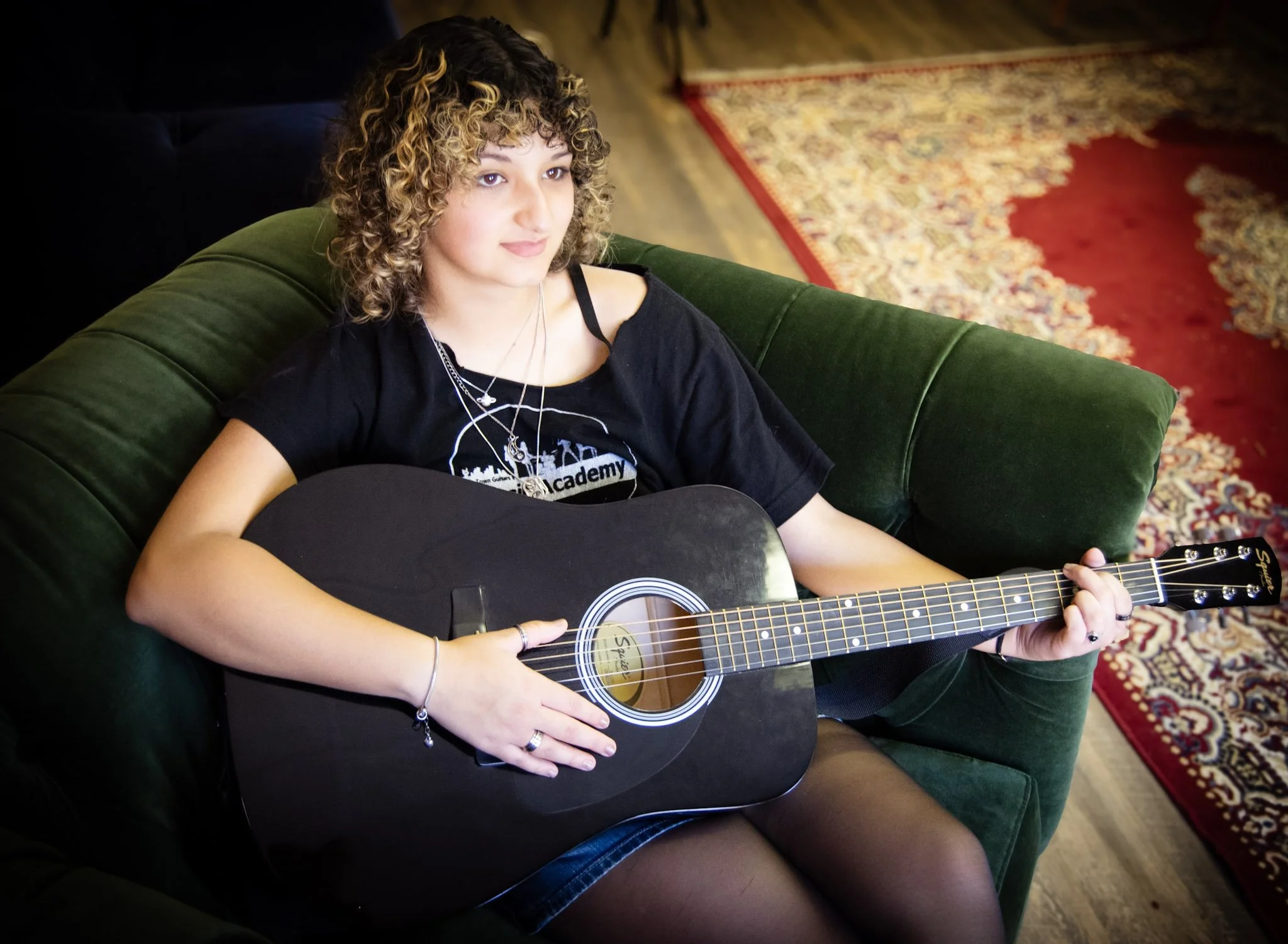 A young woman with curly hair sitting on a green velvet sofa, playing a black acoustic guitar, wearing a black T-shirt and black tights, in a room with a patterned rug.
