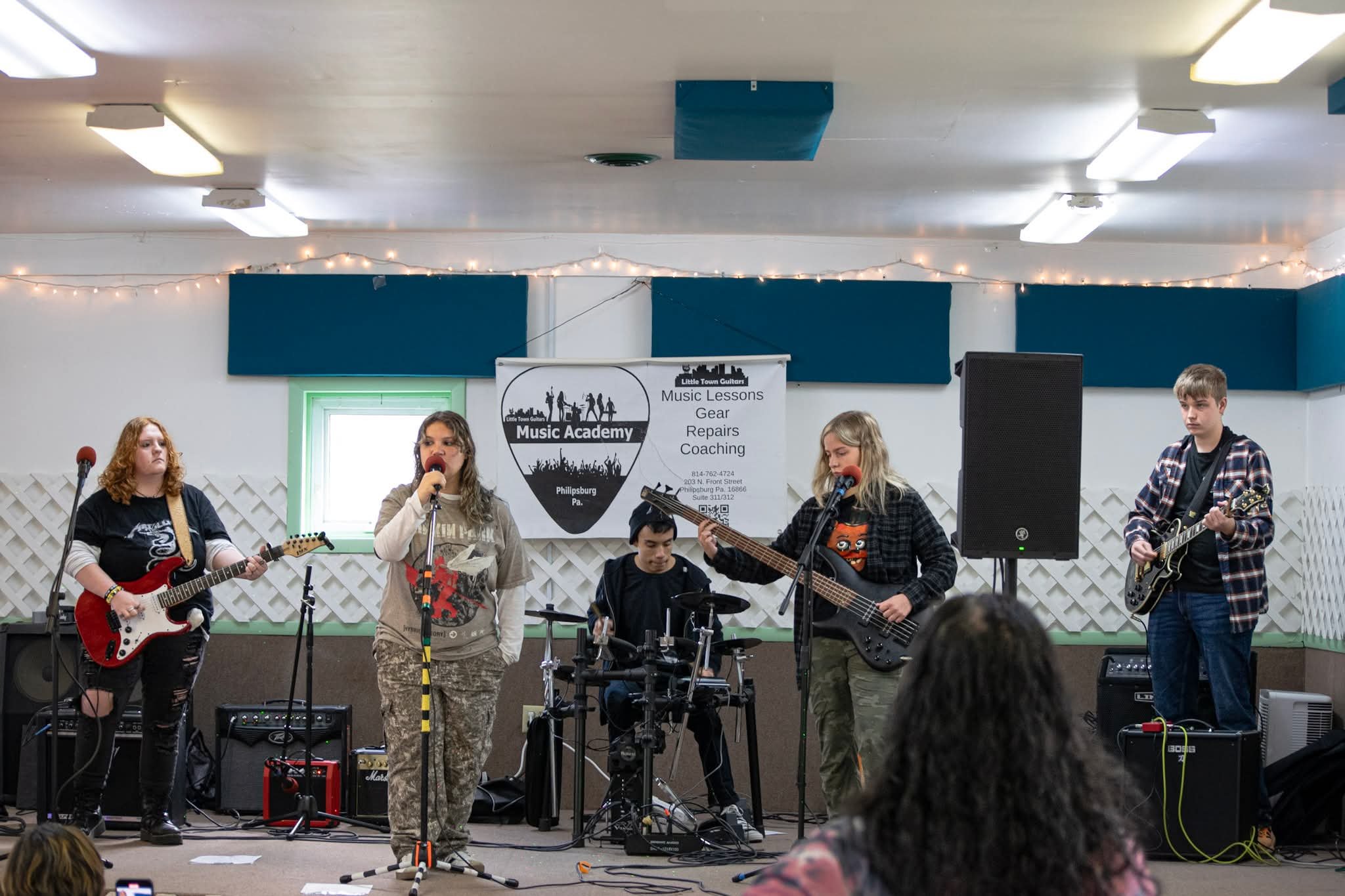 A band of five young musicians performing on a stage in an indoor setting, with a banner behind them displaying music lessons advertisement. The band includes two guitarists, a bassist, a drummer, and a female vocalist. Audience members are visible i