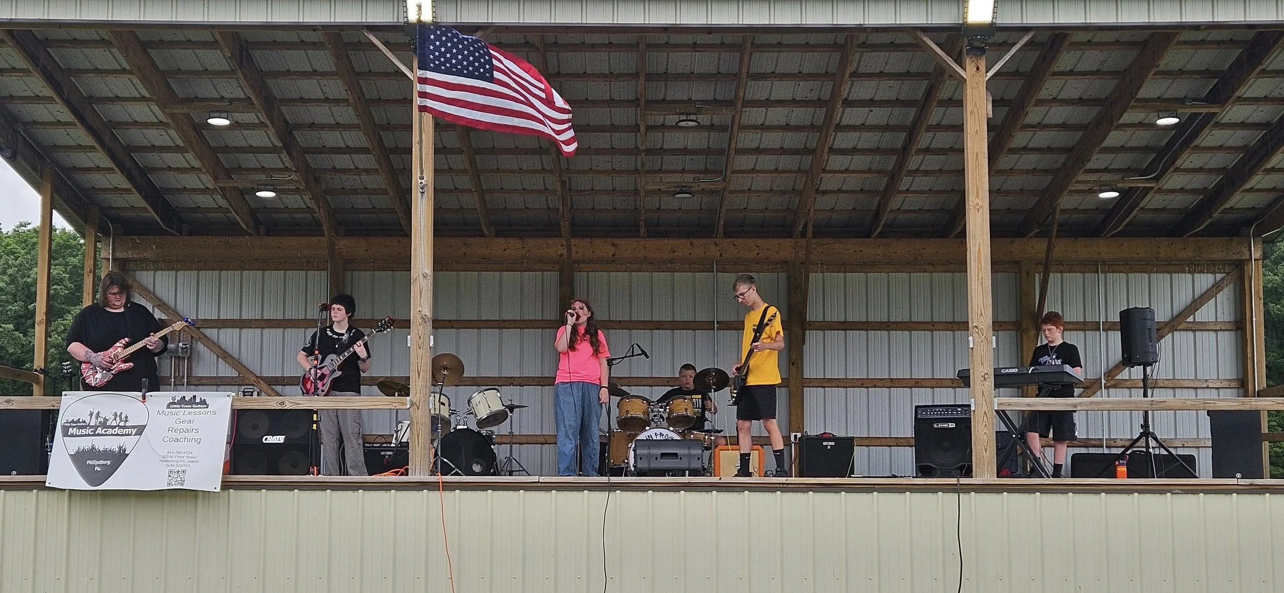A band of five young musicians performing on an outdoor stage with a wooden roof and American flag. One person is playing guitar, two are singing, one is on drums, and one is on keyboard. A sign promoting music lessons and repairs hangs on the stage.
