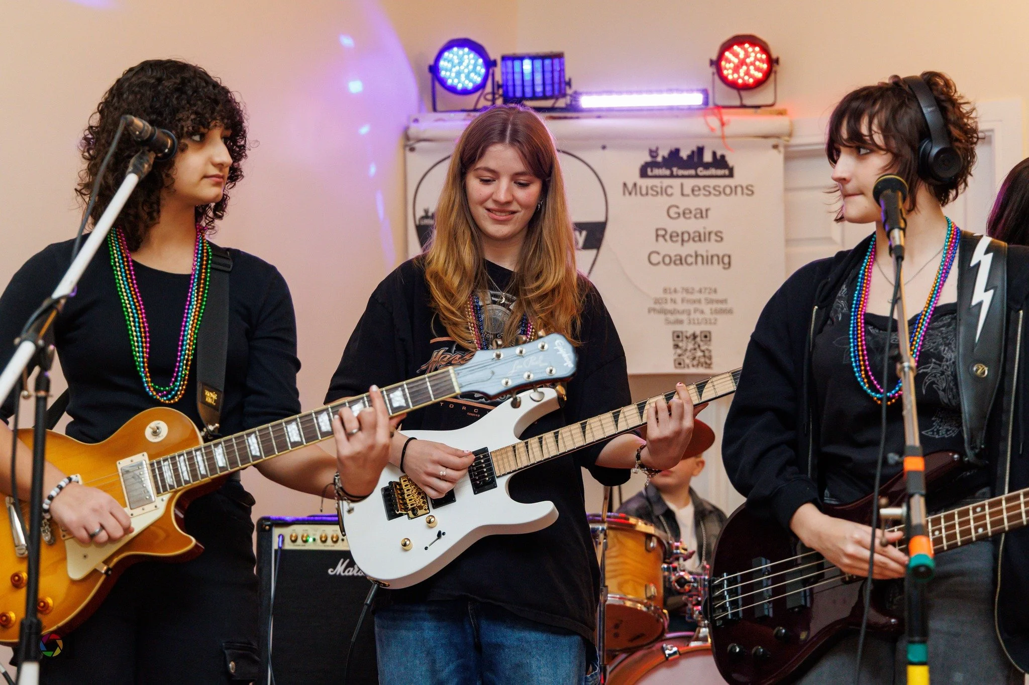 Three young women playing guitars and standing in front of a drum set at a music event, with a sign in the background displaying music lessons and services.