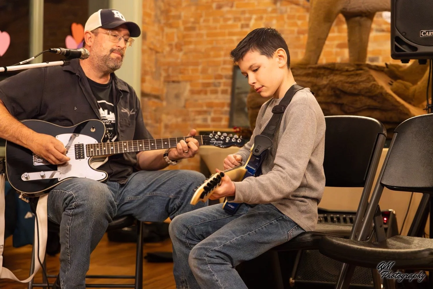 A man wearing glasses and a black shirt playing an electric guitar while sitting next to a young boy in a gray hoodie playing an electric guitar, both seated in a room with brick walls and musical equipment.