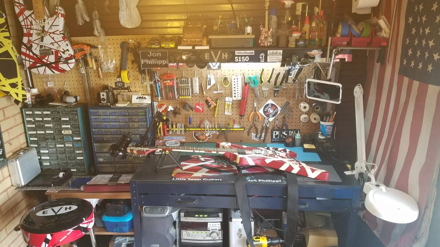 A cluttered workshop with a workbench, tools on a pegboard, and an electric guitar guitar on the workbench. There is a stool with a logo, a desk lamp, and various storage containers. The right side features an American flag curtain.